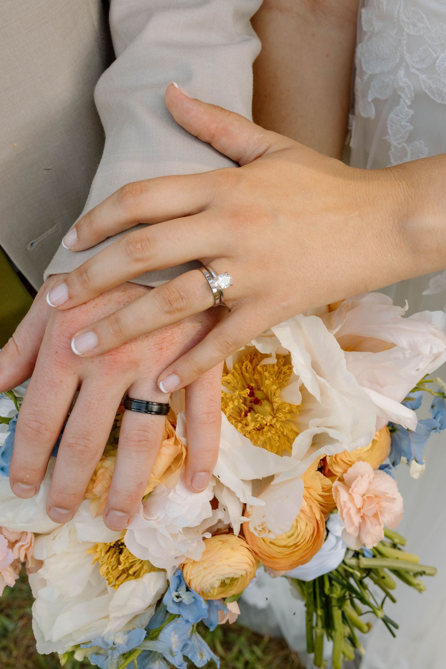 Bride and groom display wedding rings over a colorful bouquet of peach, yellow, and blue flowers.