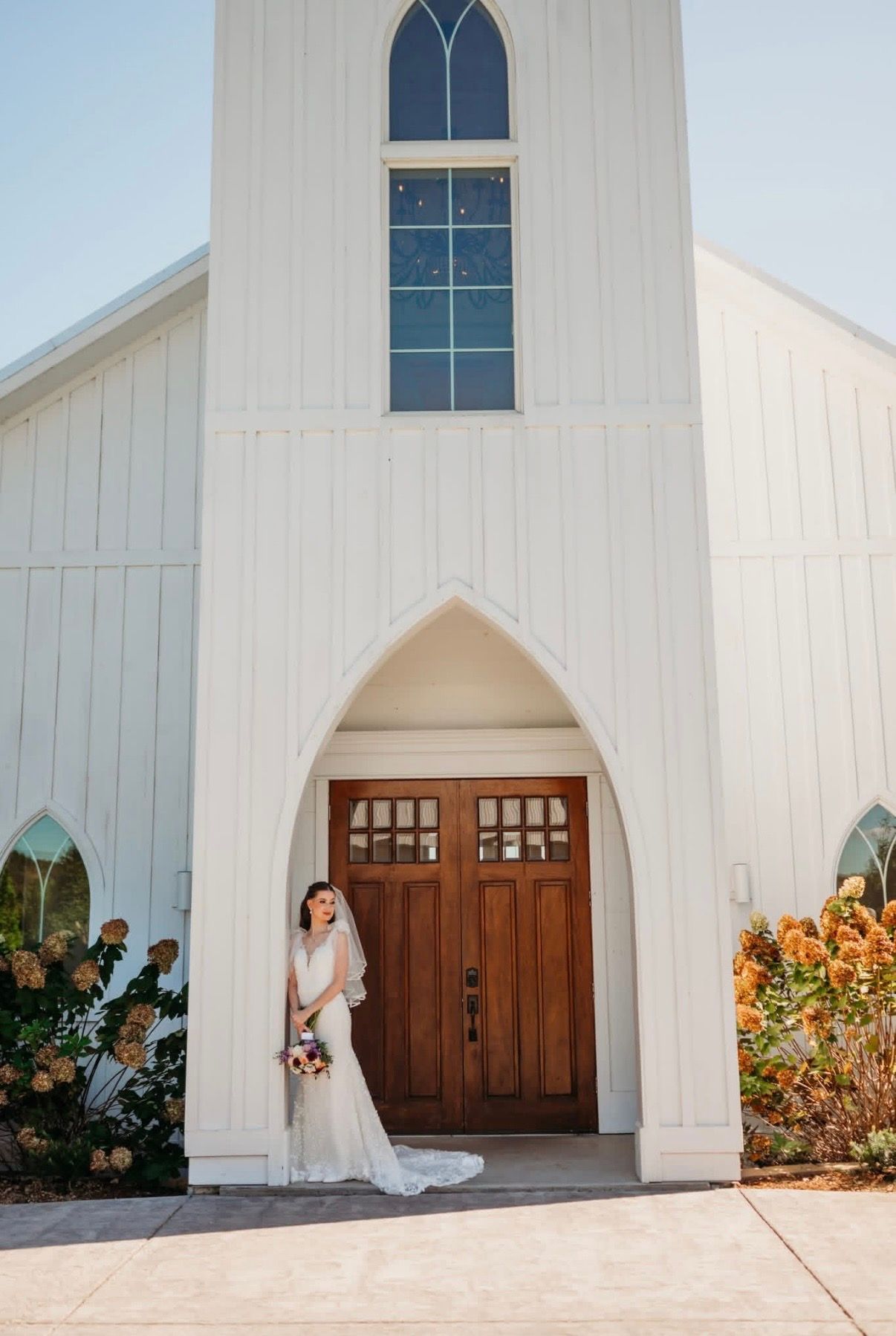 A bride in a lace gown poses at the entrance of a white chapel wedding venue.