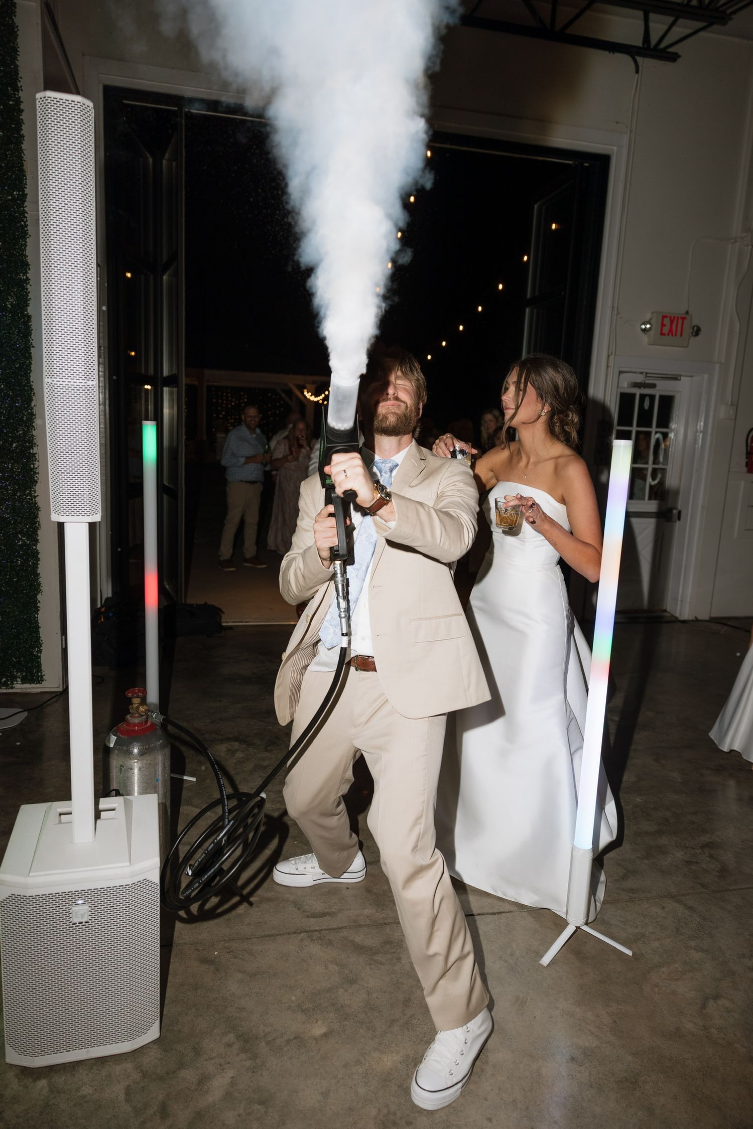 Groom in tan suit firing CO2 cannon at wedding reception while bride in white gown stands nearby.