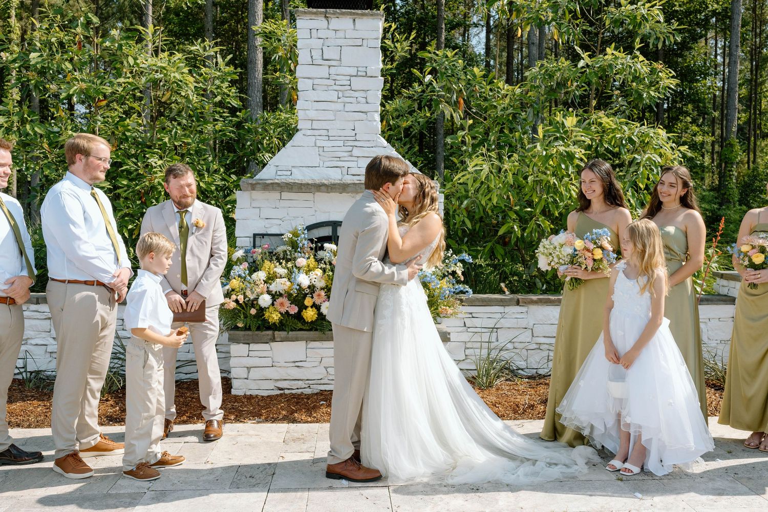 Bride and groom share first kiss at outdoor wedding ceremony with stone fireplace and wedding party beside them.