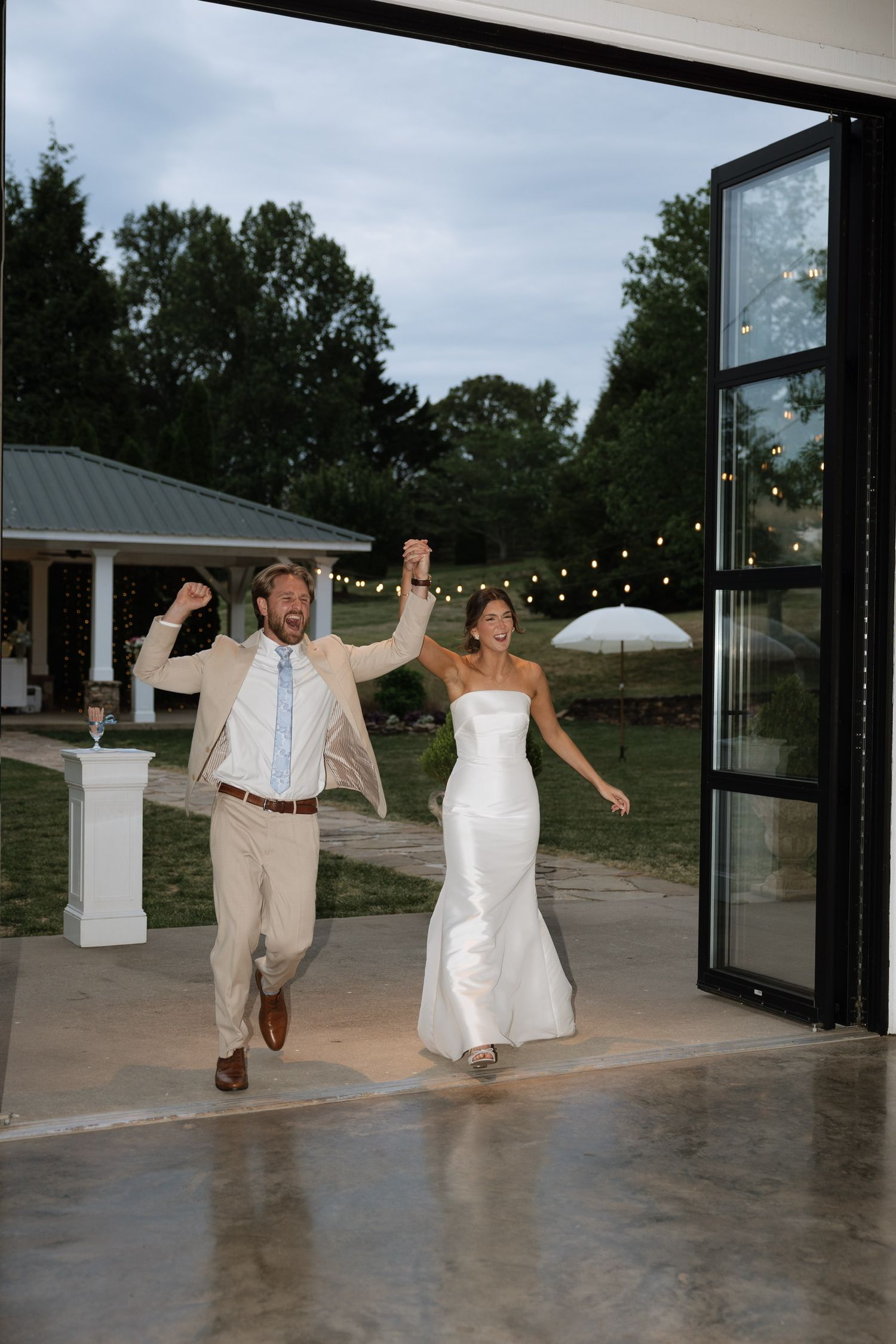 Bride in white gown and groom in tan suit joyfully enter wedding reception through glass doors at dusk.