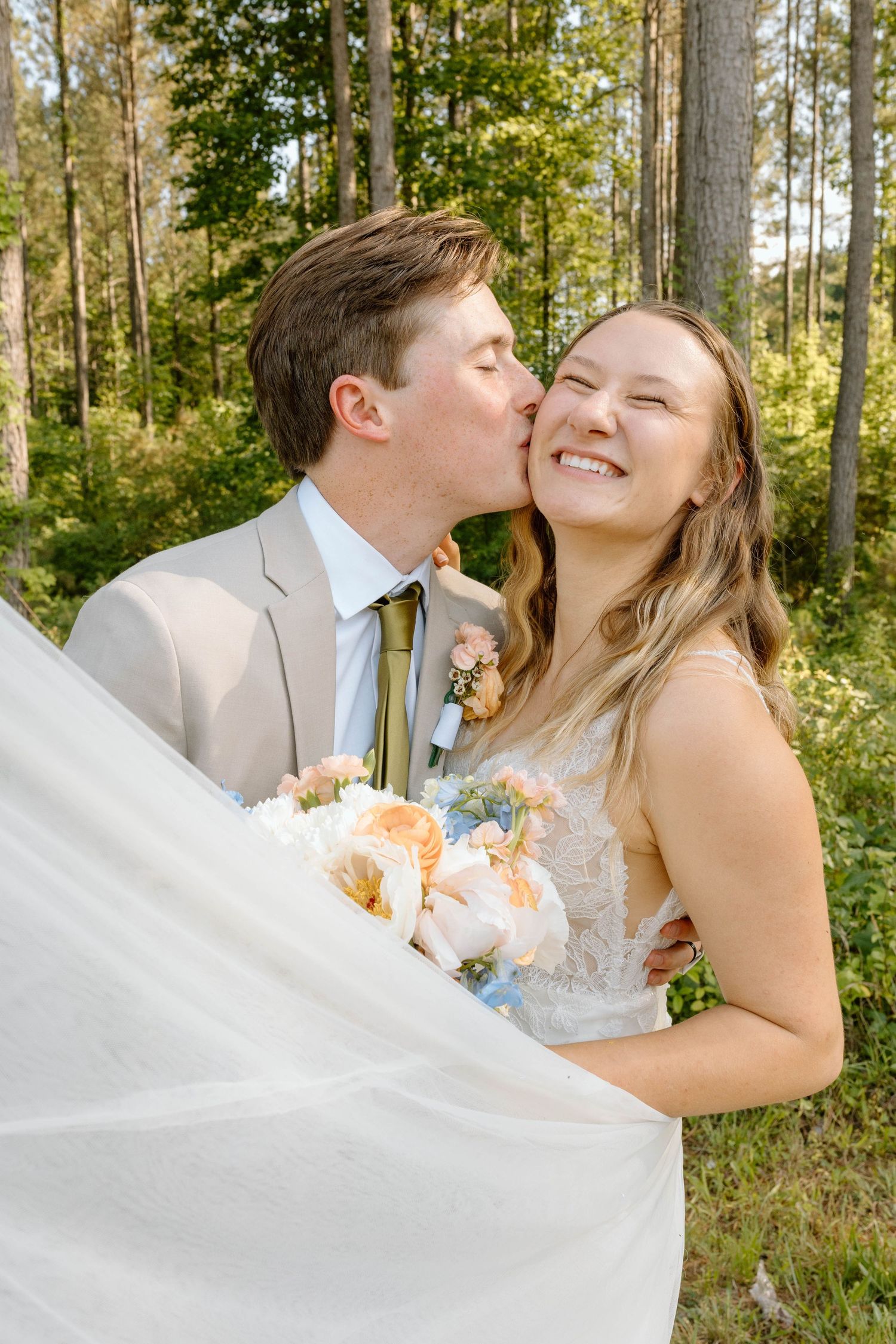 Smiling bride holding peach bouquet gets kissed on cheek by groom in tan suit outdoors in forest setting.