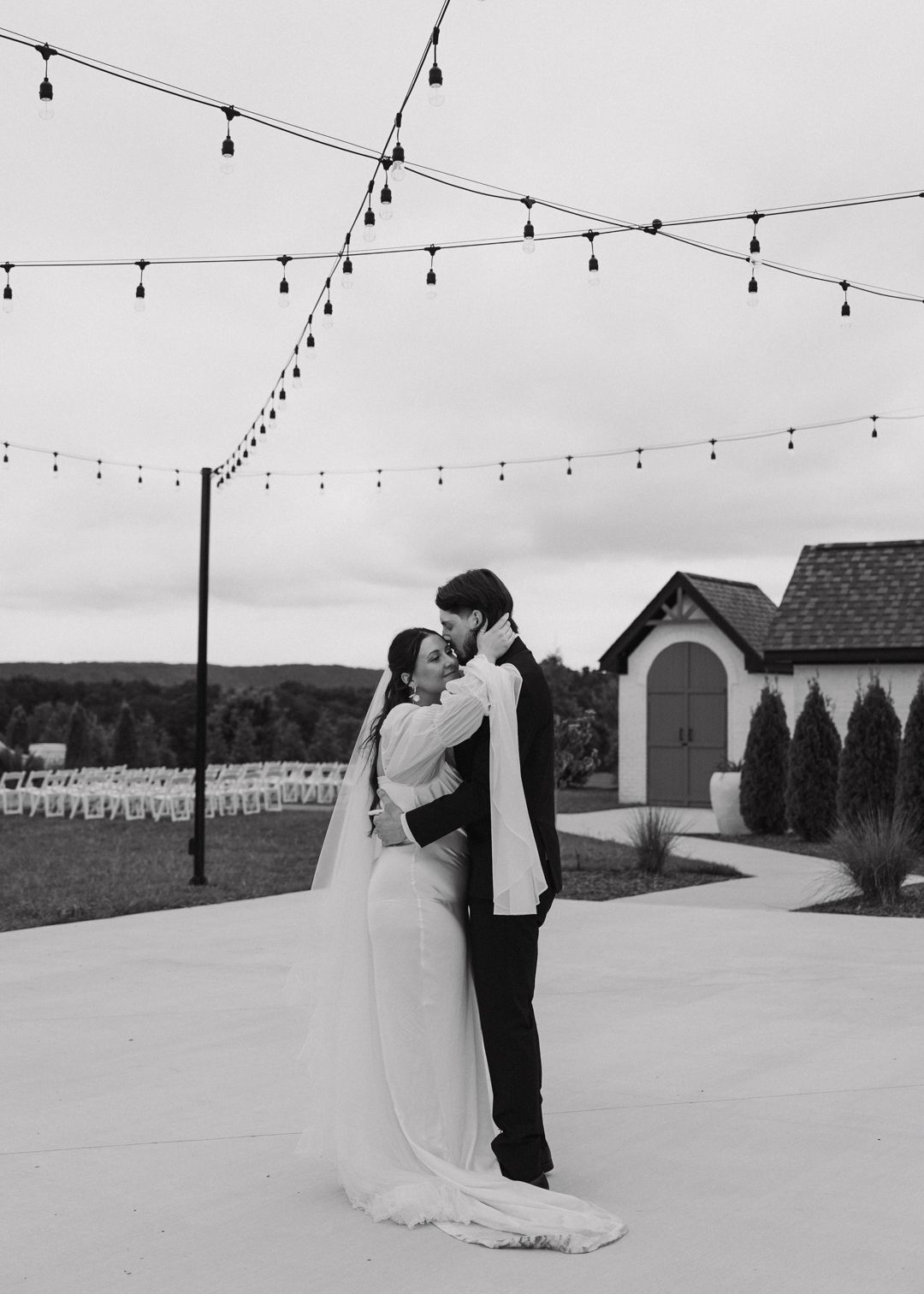 Black and white photo of bride and groom embracing under string lights at outdoor wedding venue.