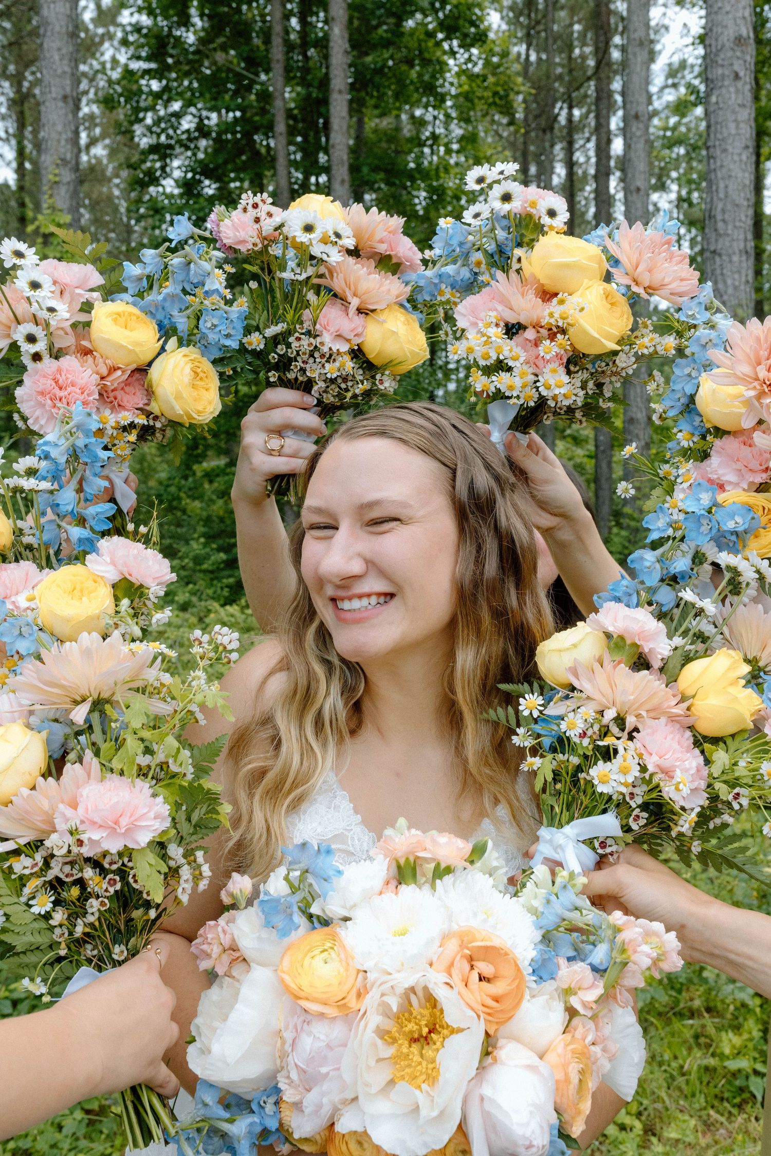 Smiling bride framed by colorful floral wreath of yellow, pink, and blue flowers in a forest setting.