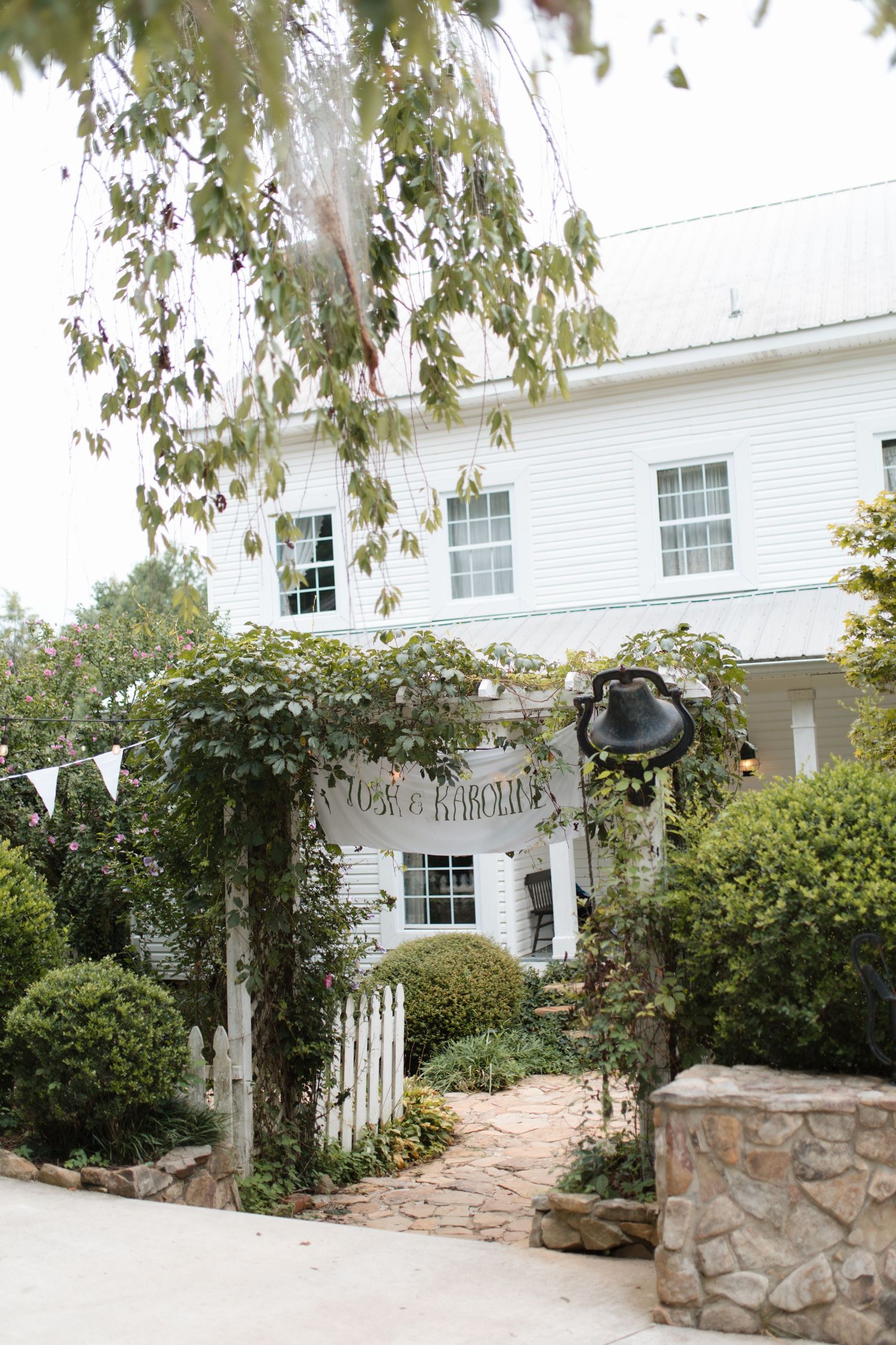 Charming white farmhouse wedding venue with lush greenery arbor entrance, brick pathway, and decorative bunting flags.