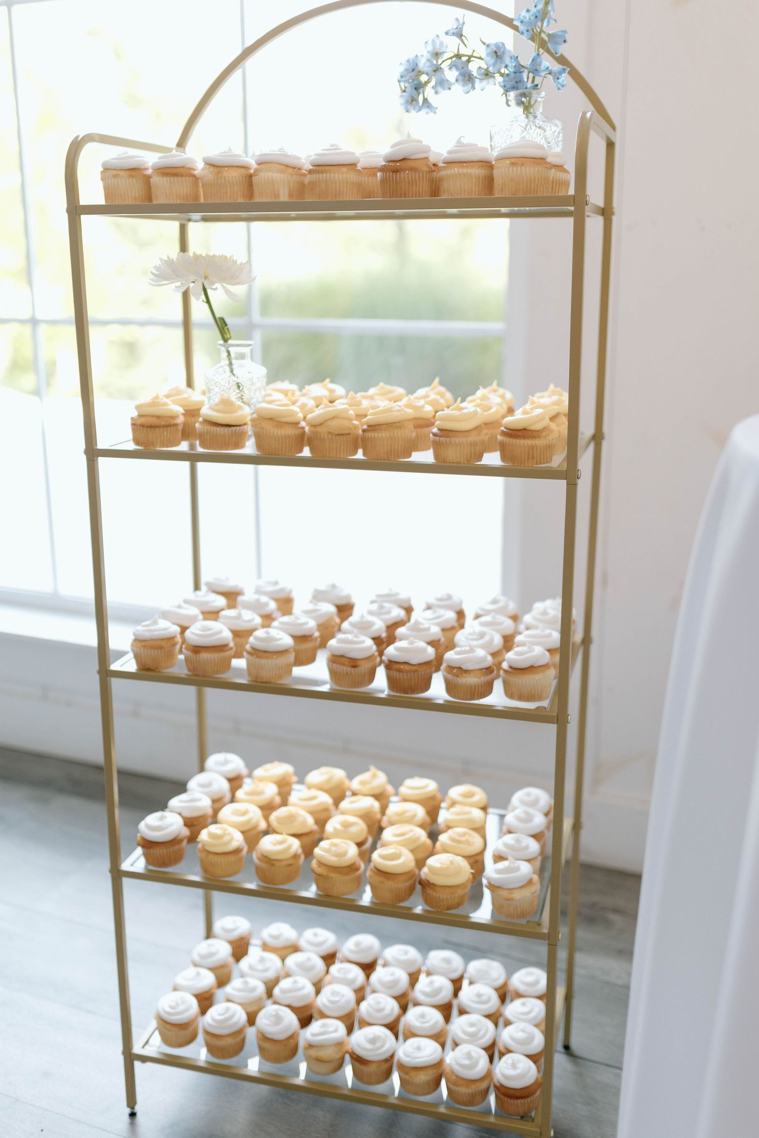 Elegant gold tiered display shelf filled with rows of frosted vanilla cupcakes at a wedding reception.