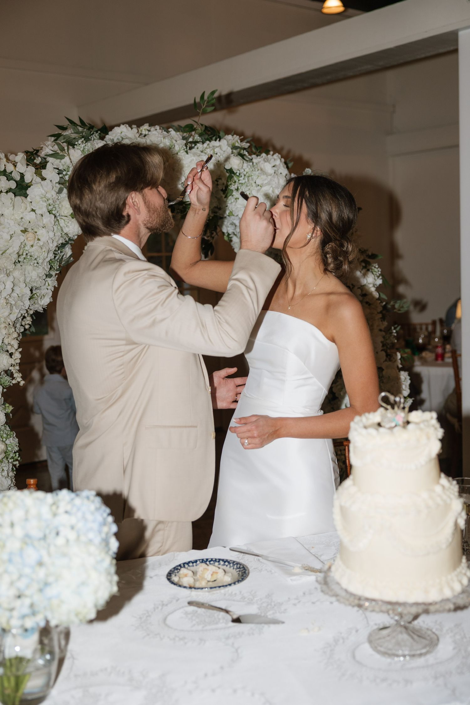 Bride and groom share cake cutting moment beneath white floral arch at wedding reception.