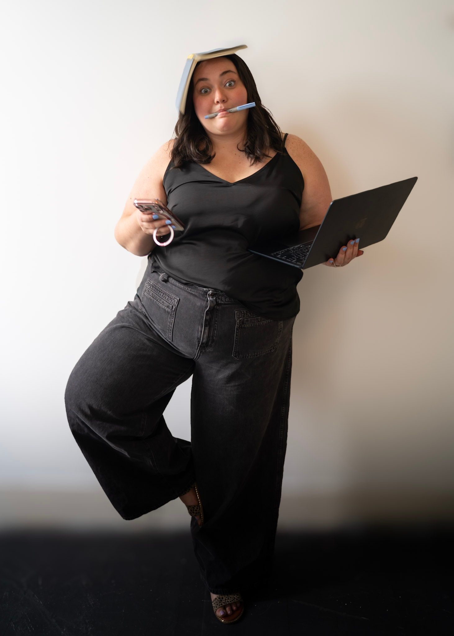 Woman balancing book on head, holding laptop and phone, pen in mouth, wearing black outfit, multitasking pose.