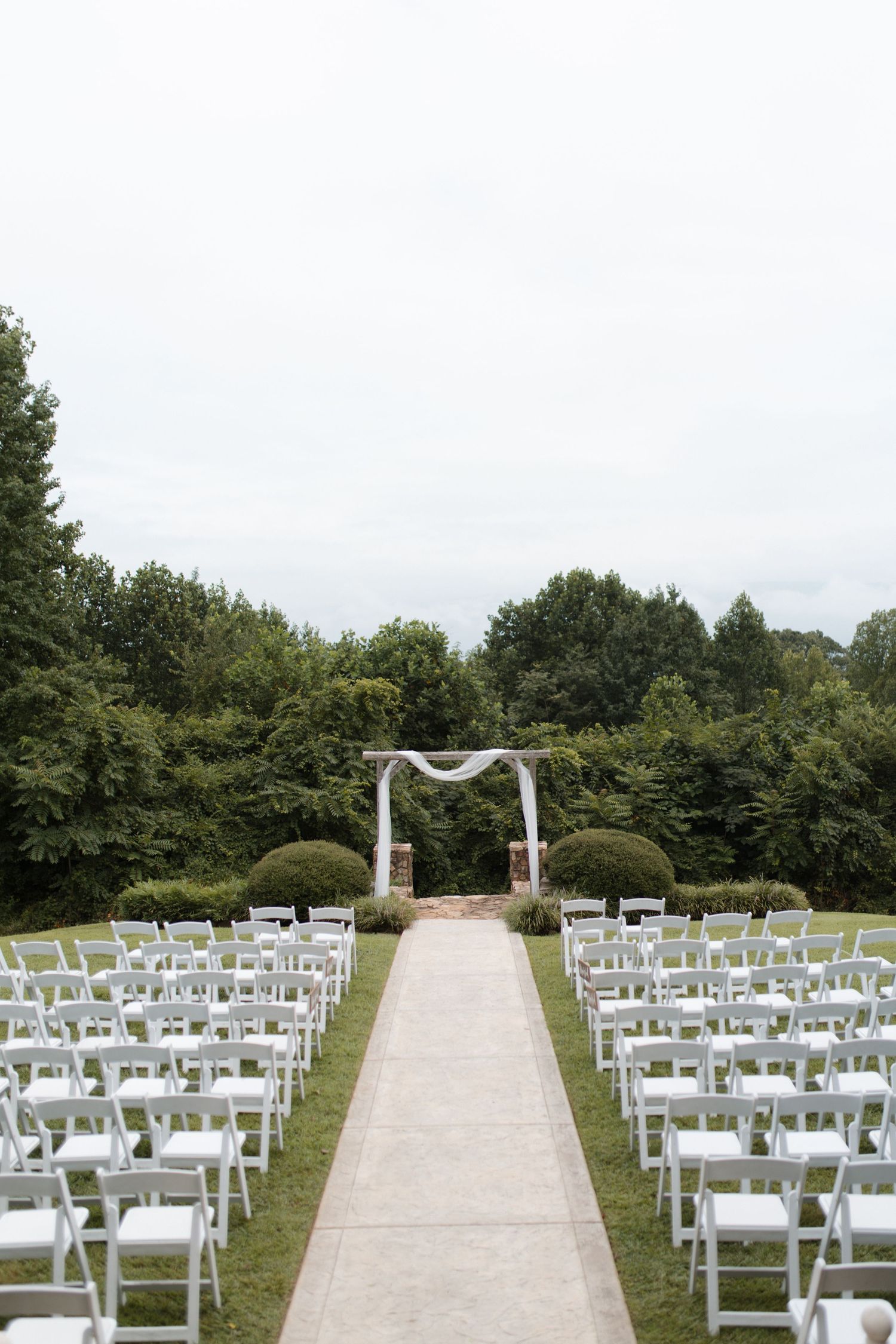 Outdoor wedding ceremony setup with white chairs lining aisle leading to draped floral arch.