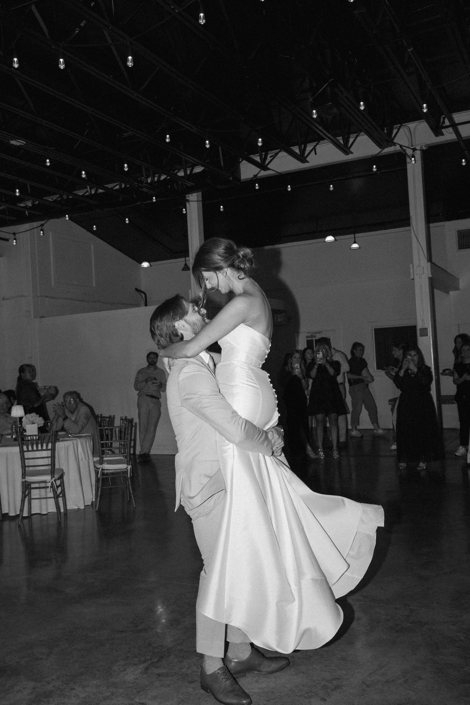 Black and white photo of groom lifting bride during first dance at elegant wedding reception venue.