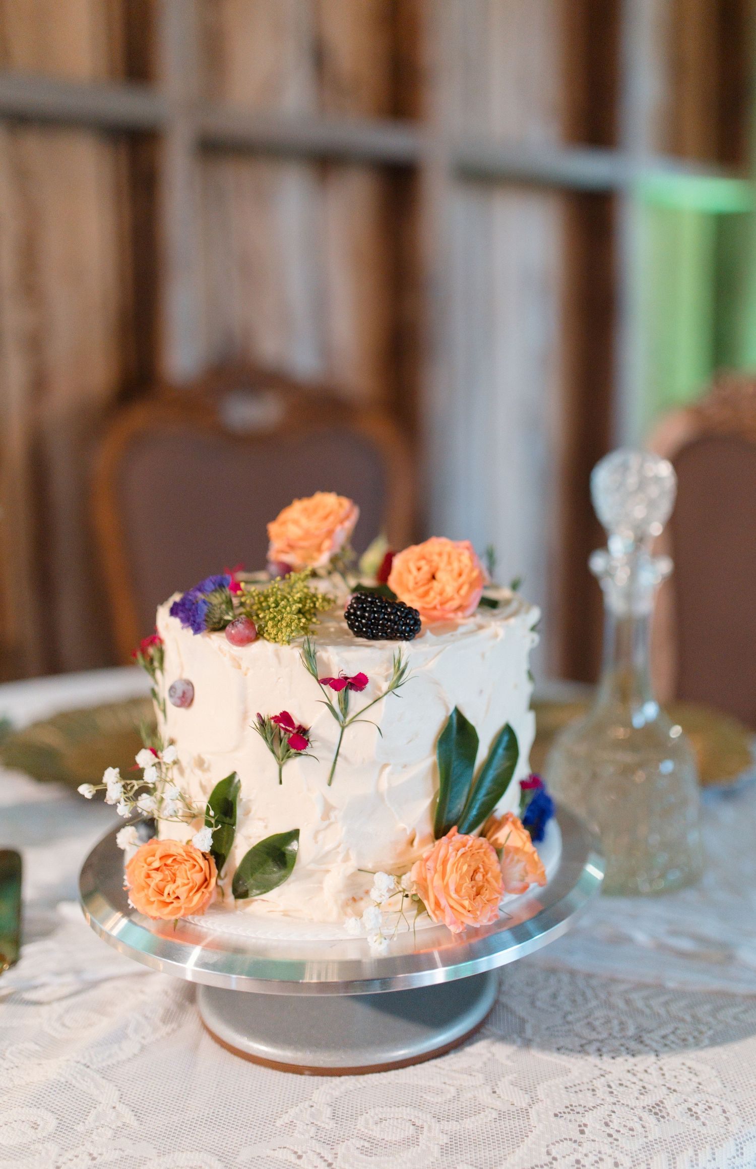 Elegant single-tier white wedding cake decorated with fresh peach roses, blackberries, and colorful pressed flowers on stand.