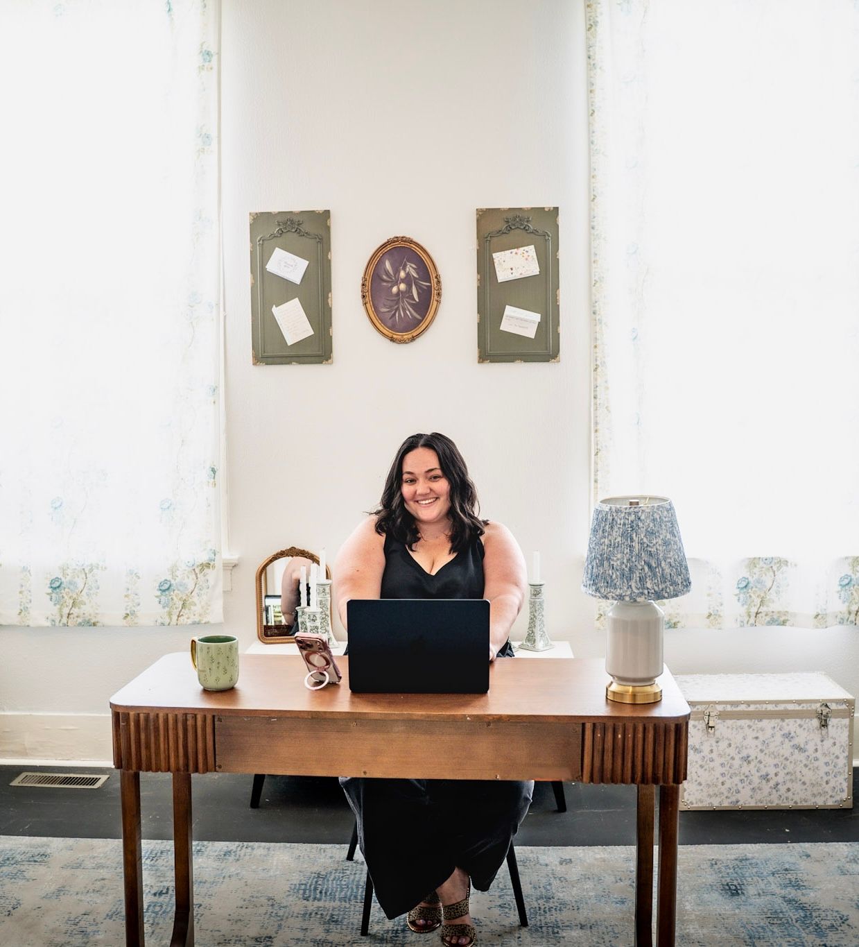 Smiling wedding planner sits at wooden desk with laptop in bright, elegantly decorated office.