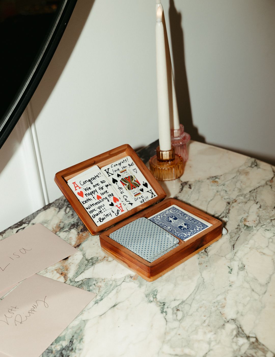 Wooden playing card box with handwritten notes open on marble table beside taper candle.
