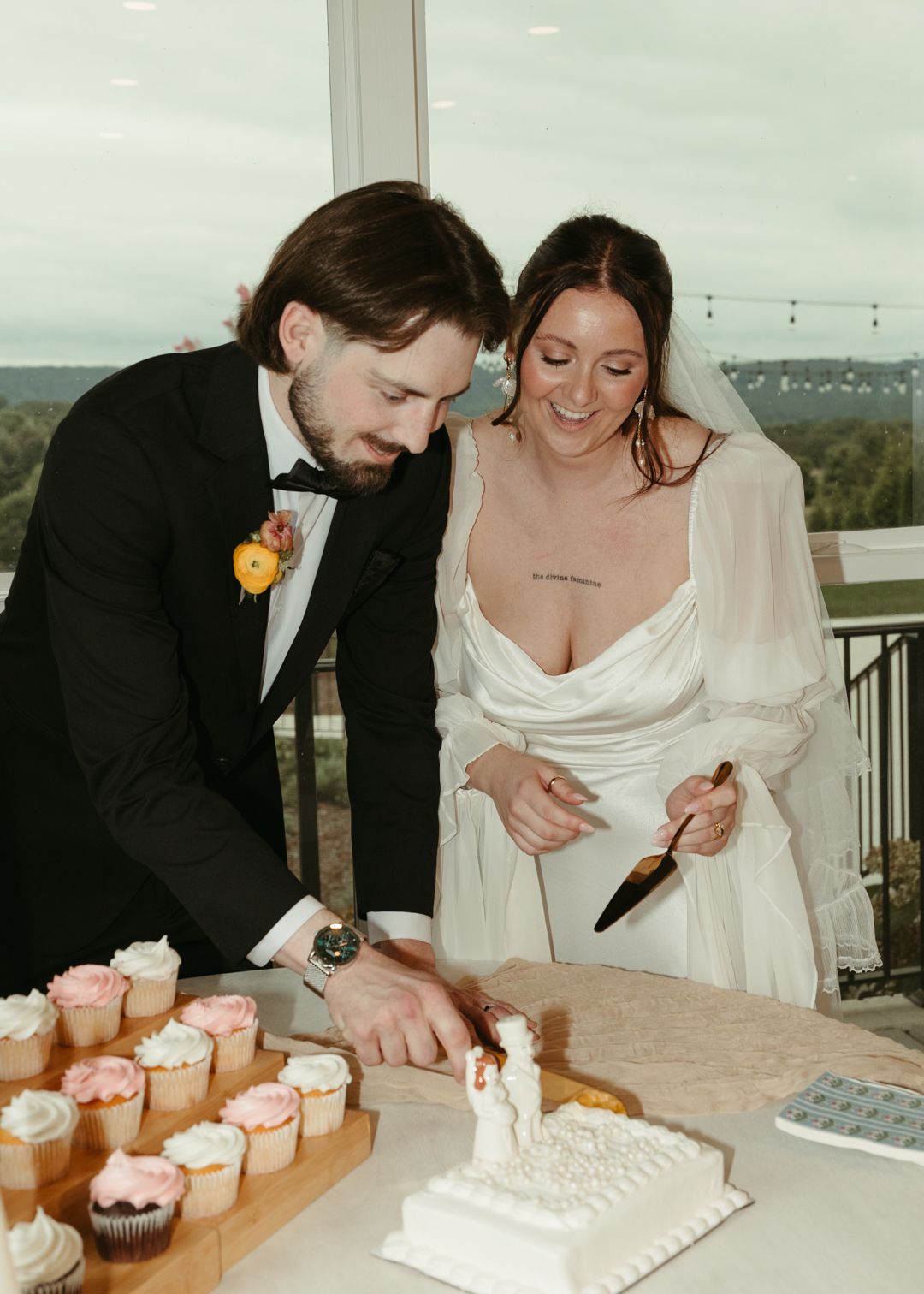 Smiling bride and groom cutting white wedding cake surrounded by pink and white cupcakes.
