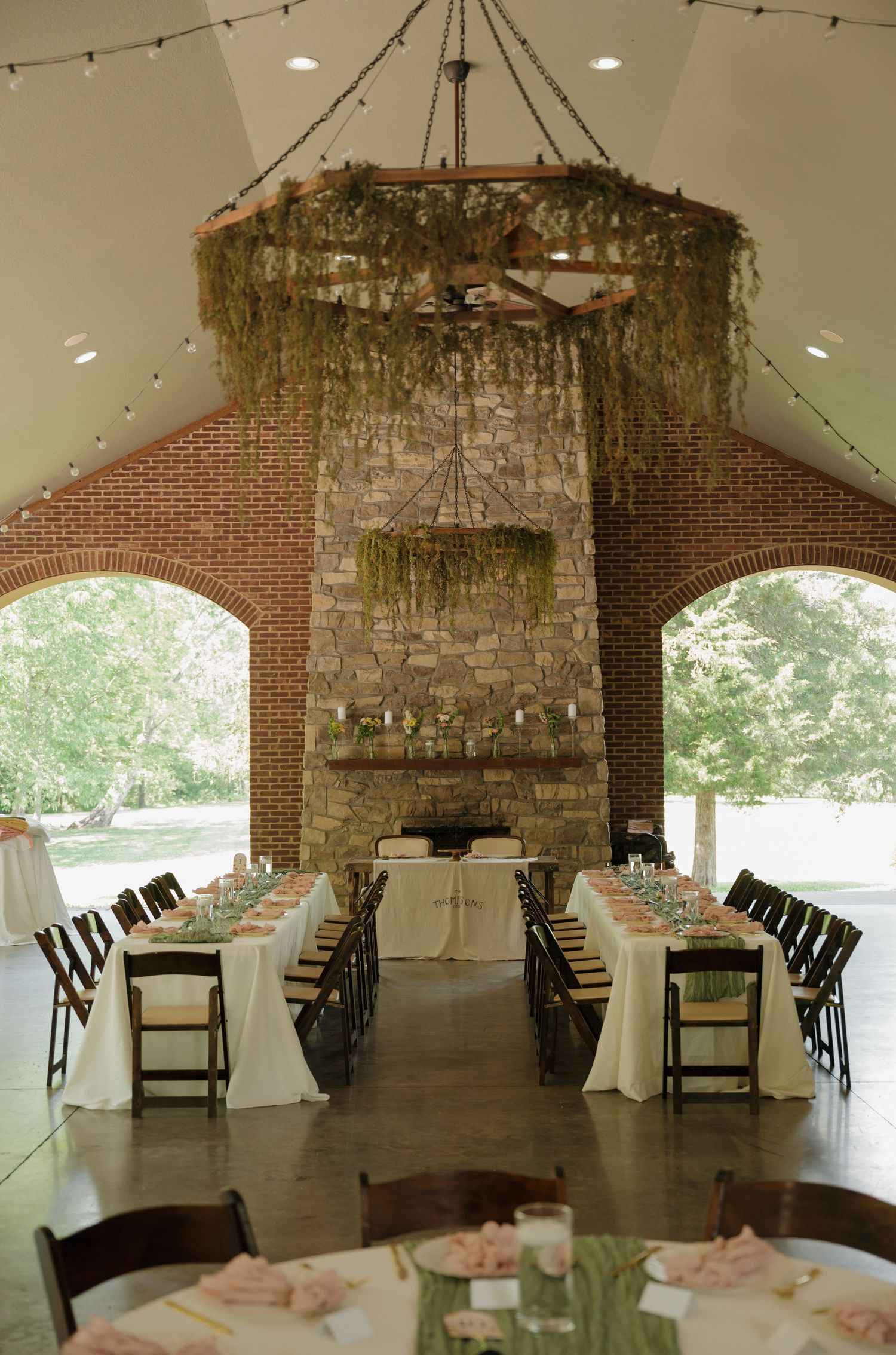 Elegant wedding reception hall with brick fireplace, hanging greenery chandelier, and long decorated dining tables.