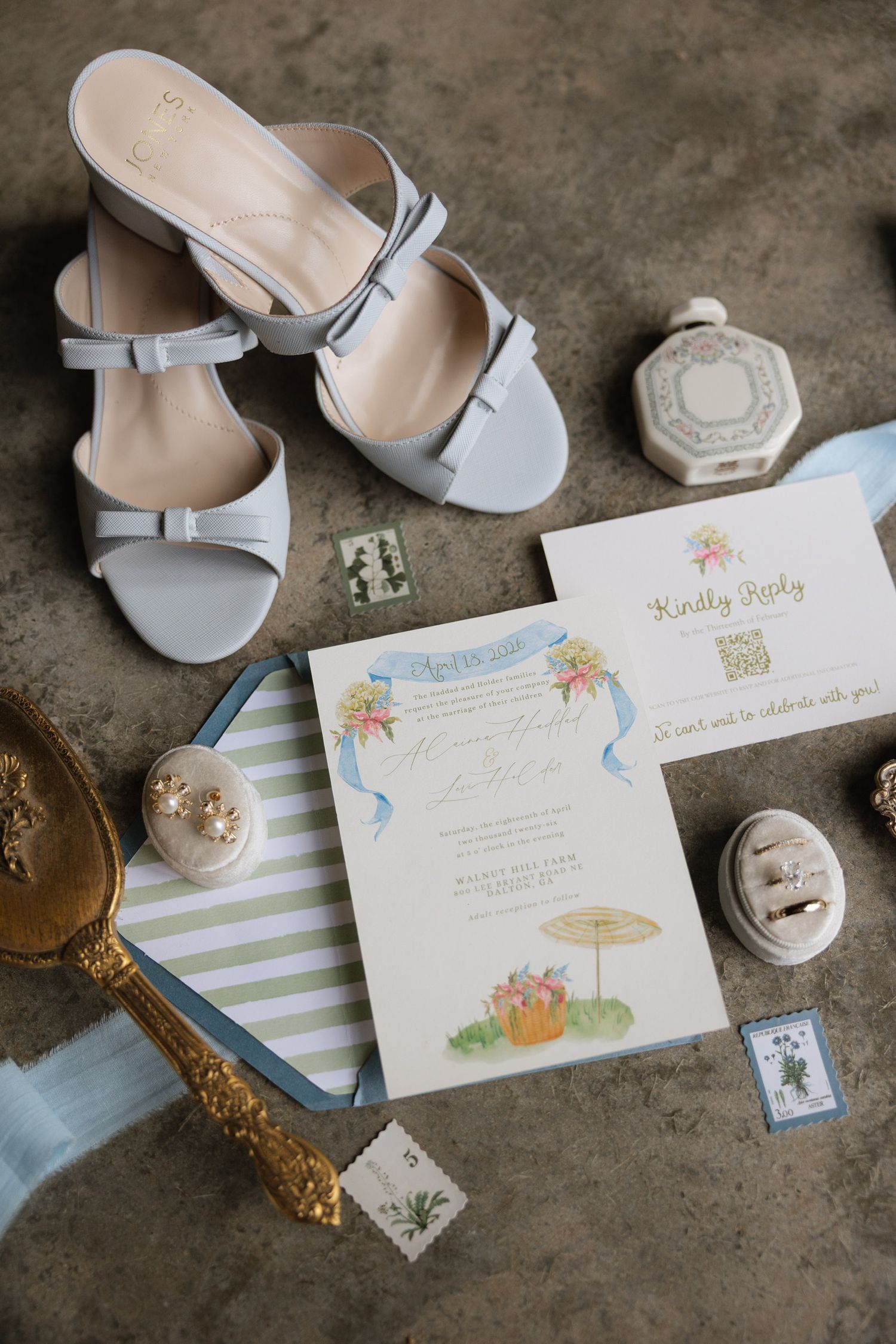 Wedding flat lay with blue heels, floral invitation suite, reply card, stamps, and ring boxes.