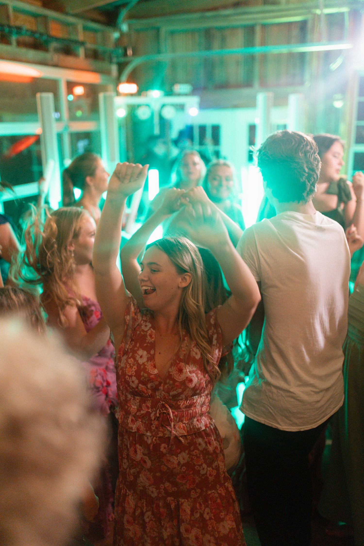 Guests dancing energetically under green lights at vibrant wedding reception, woman in floral dress with arms raised.