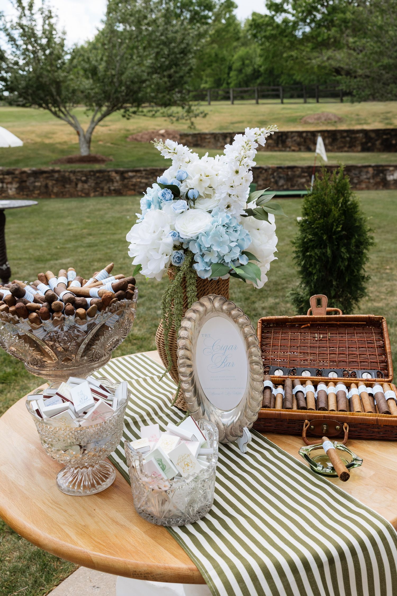 Outdoor wedding favor table with cigars, matchboxes, floral arrangement, and green striped runner.