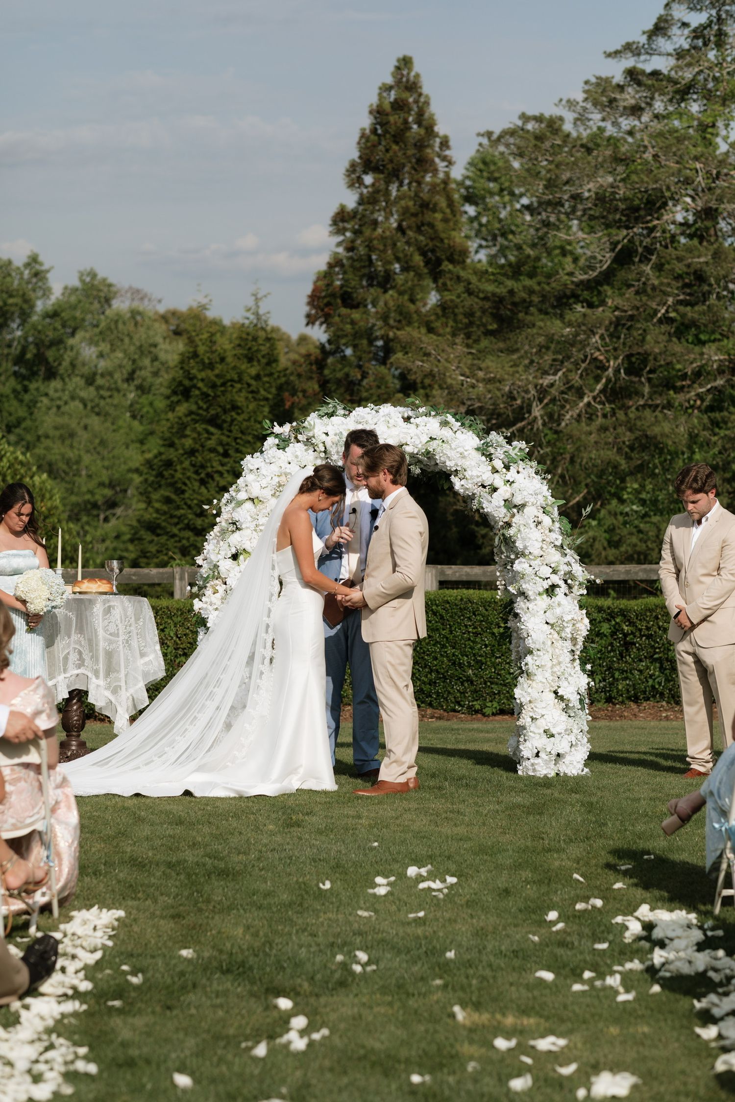 Outdoor wedding ceremony with bride and groom under white floral arch on lush green lawn with guests seated nearby.