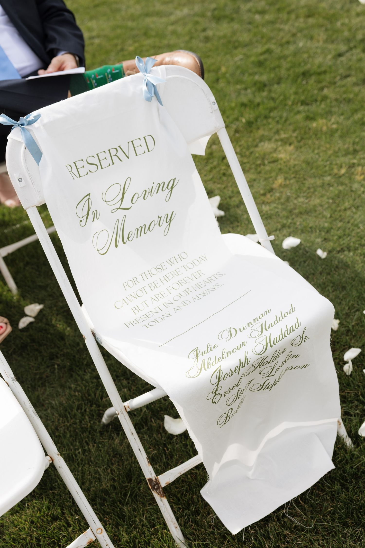 White wedding chair with In Loving Memory reserved sign featuring gold script and blue ribbon bow.