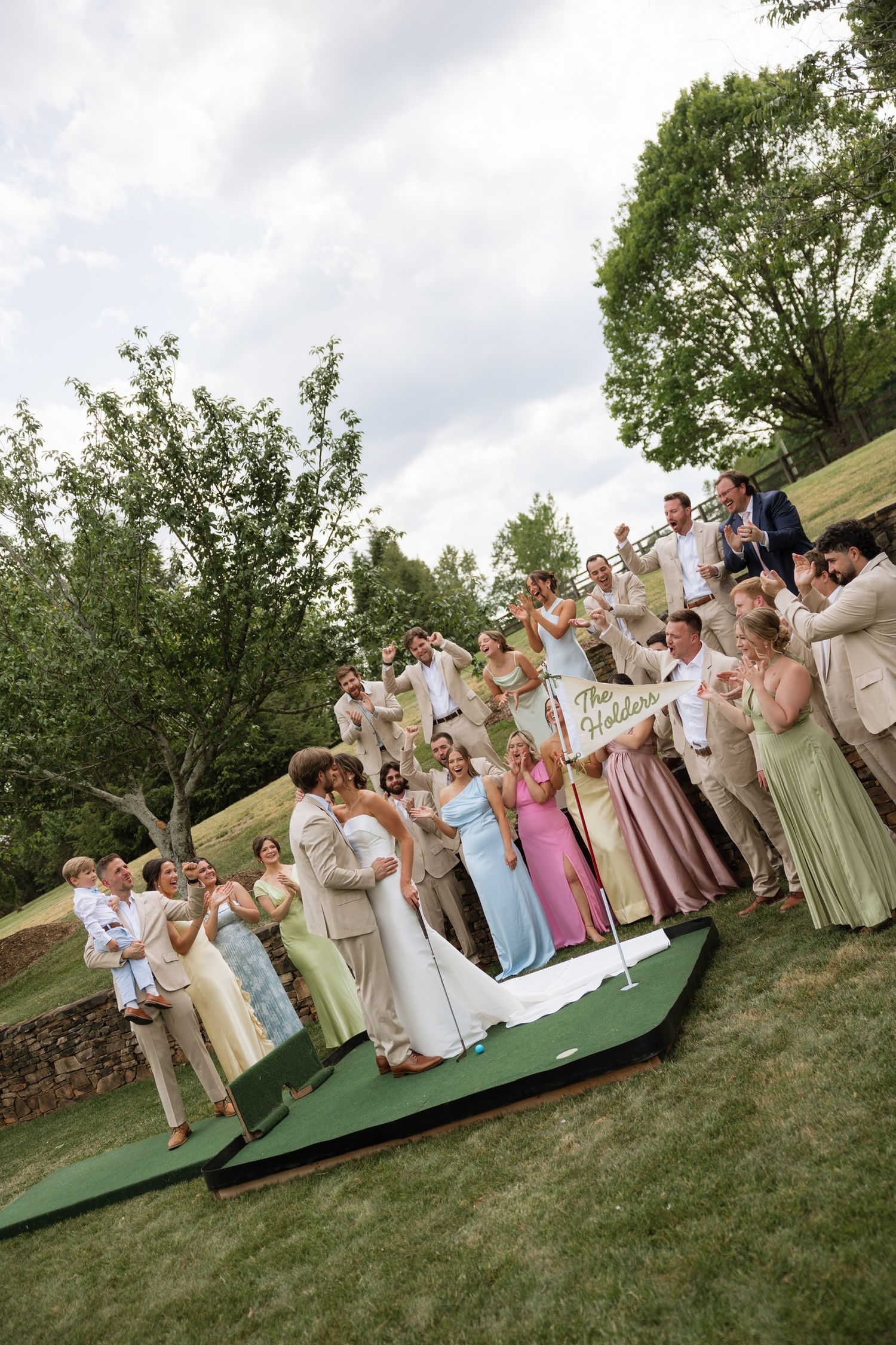 Wedding party and guests cheering outdoors near a putting green with a newlywed sign.