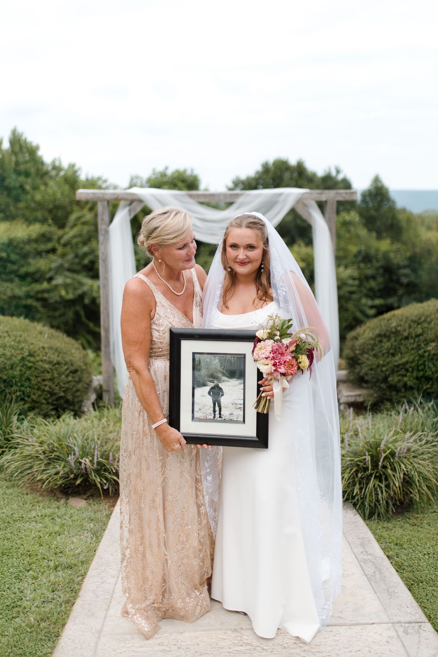 Bride in white gown and veil holds bouquet with mother in gold dress, displaying framed photo at outdoor ceremony.