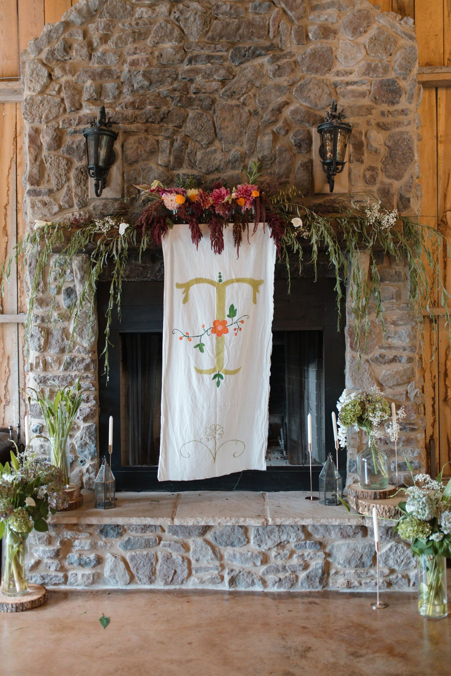 Rustic stone fireplace decorated with monogram banner, cascading greenery, and colorful floral arrangements for wedding reception.