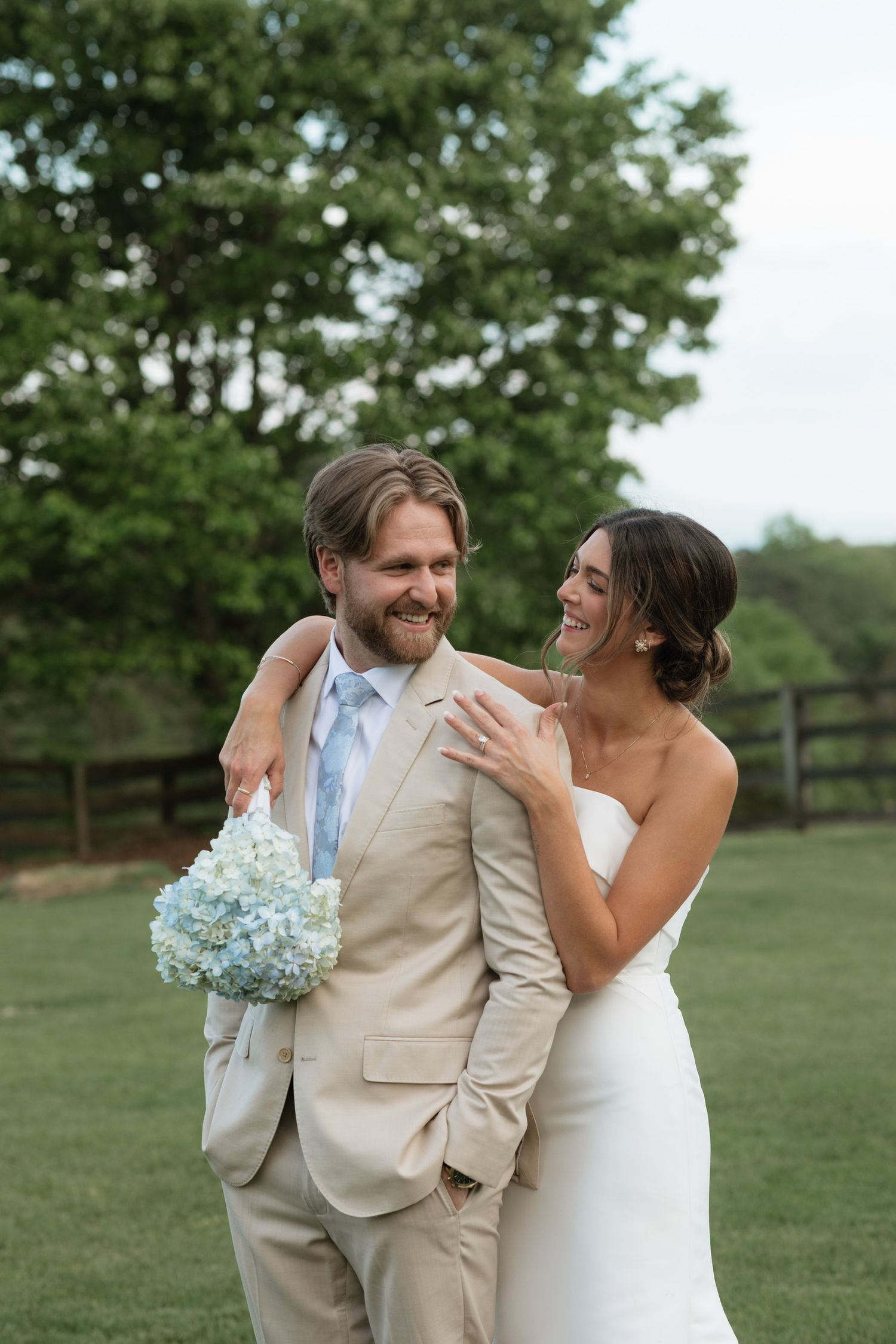 A smiling bride in a white strapless gown embraces a bearded groom in a tan suit holding a blue hydrangea bouquet outdoors.