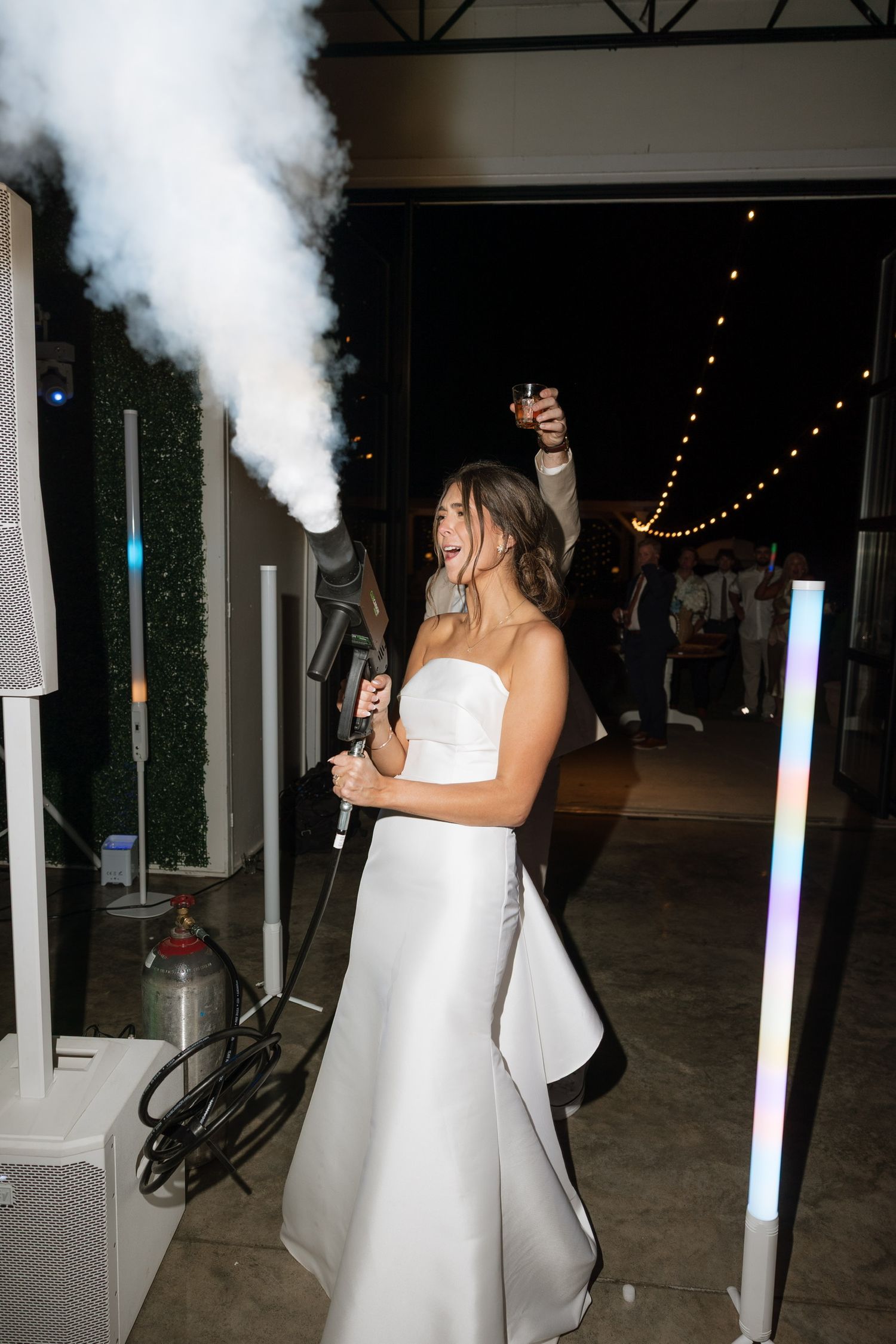 Bride in strapless white gown laughing while firing CO2 cannon at evening wedding reception.