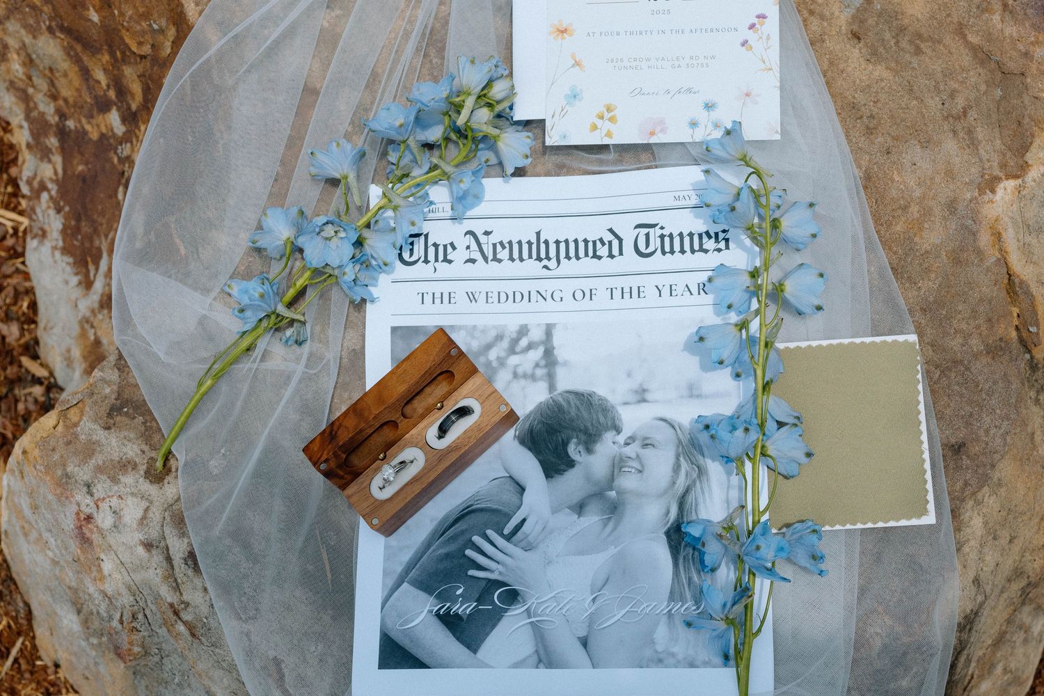 Wedding stationery flat lay featuring The Newlywed Times newspaper, rings, veil, and blue floral garland on stone.
