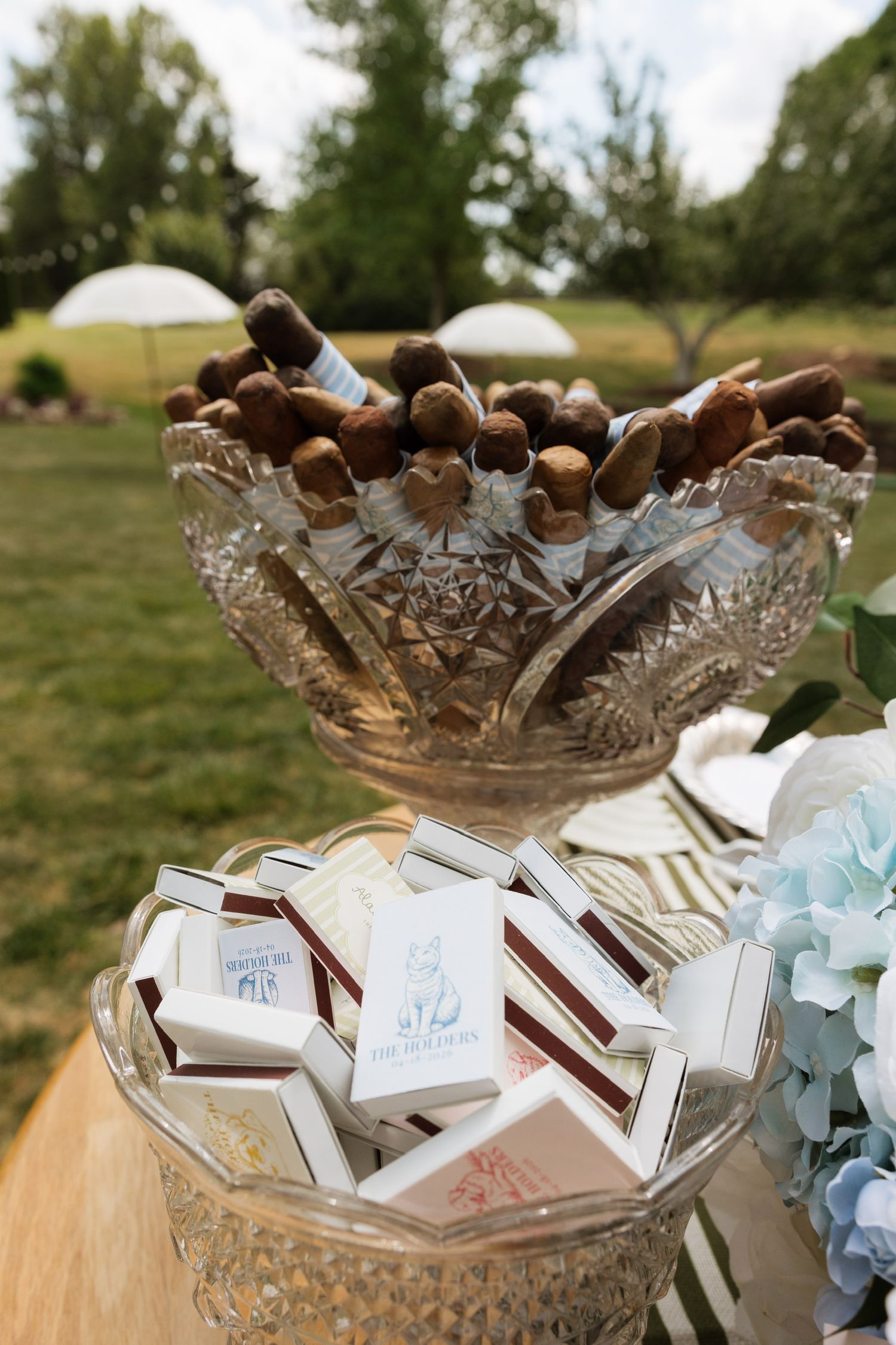 Crystal bowls filled with cigars and custom matchbox wedding favors displayed outdoors.