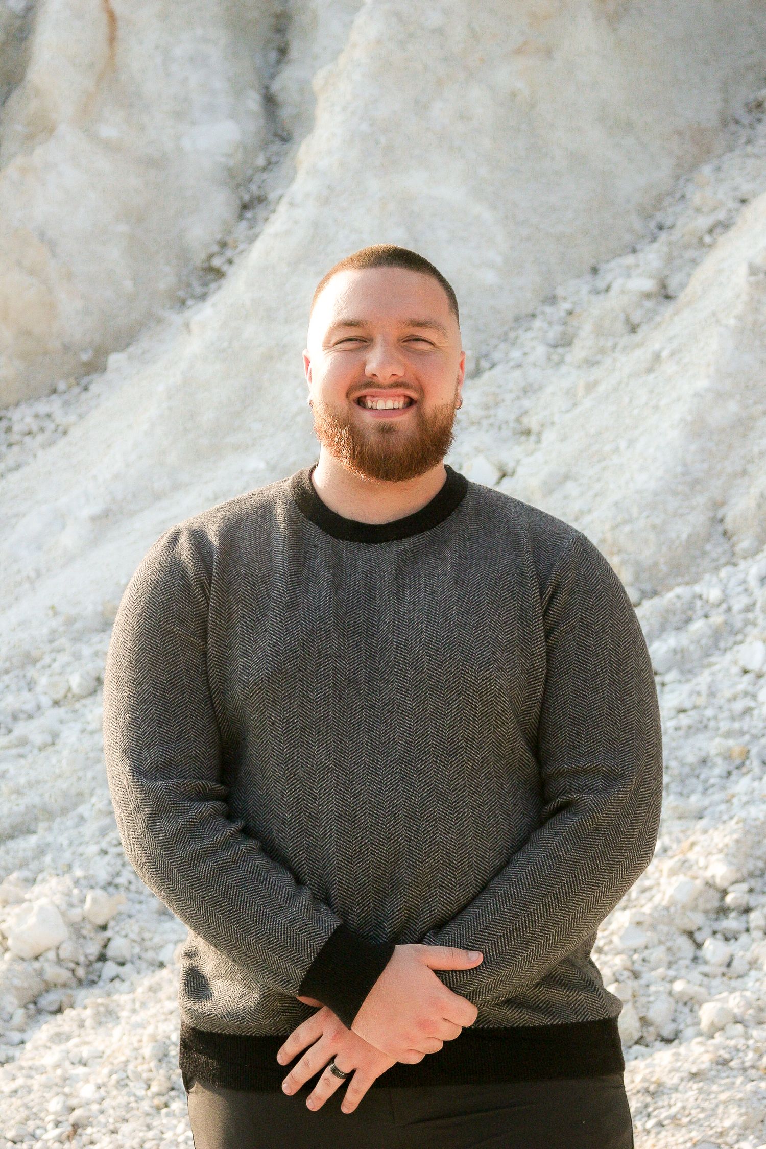 Smiling bearded man in gray sweater standing outdoors against white rocky cliff background.