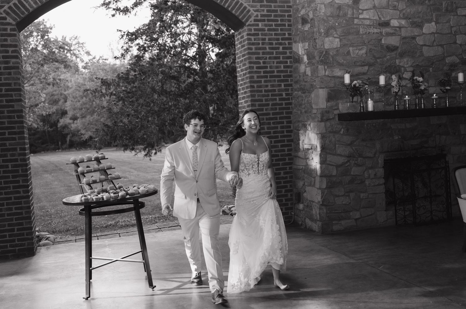 Bride and groom walking hand in hand through brick archway at outdoor wedding venue in black and white photo.