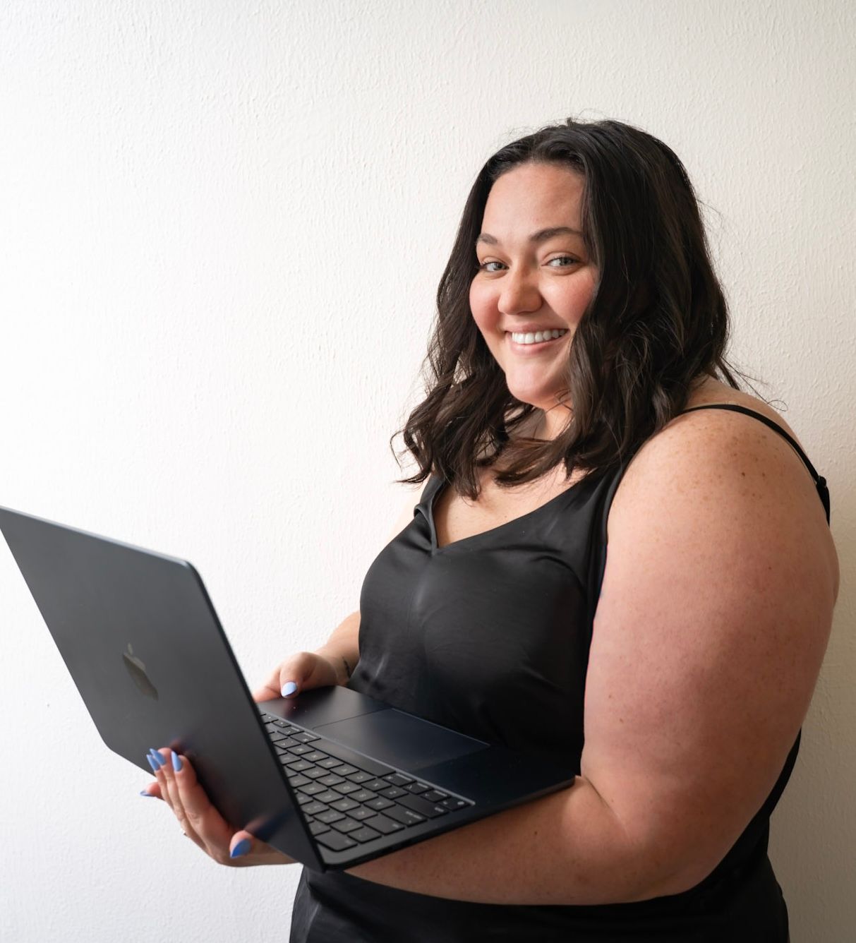 Smiling woman holding a laptop computer while posing against a white wall indoors.