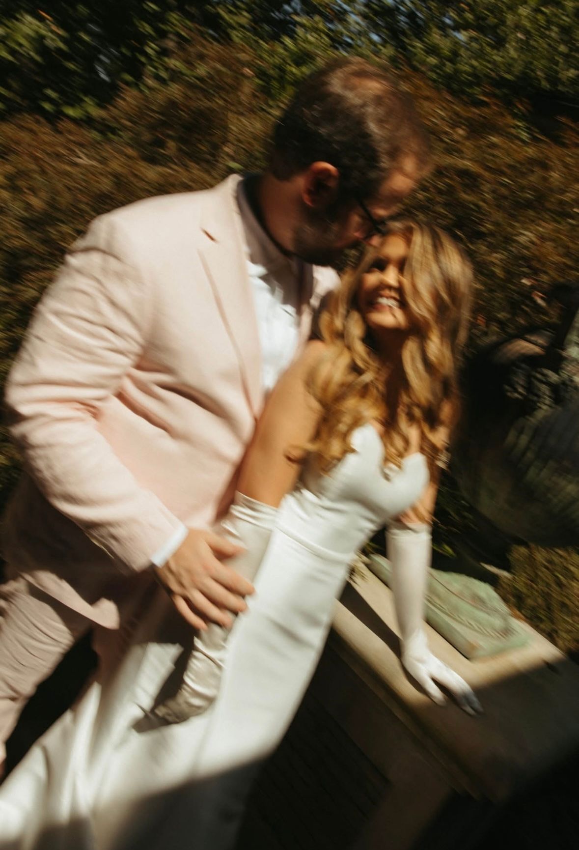 Groom in blush pink suit embracing smiling bride in white gown in a dreamy motion-blur outdoor photo.