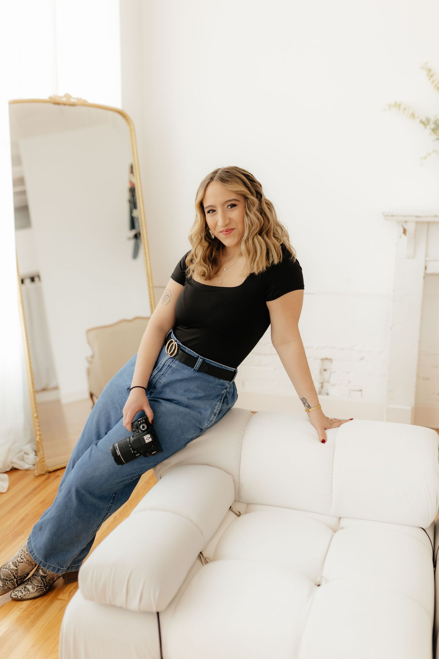 Blonde woman in black top and jeans holding camera while sitting on white sofa near gold mirror.