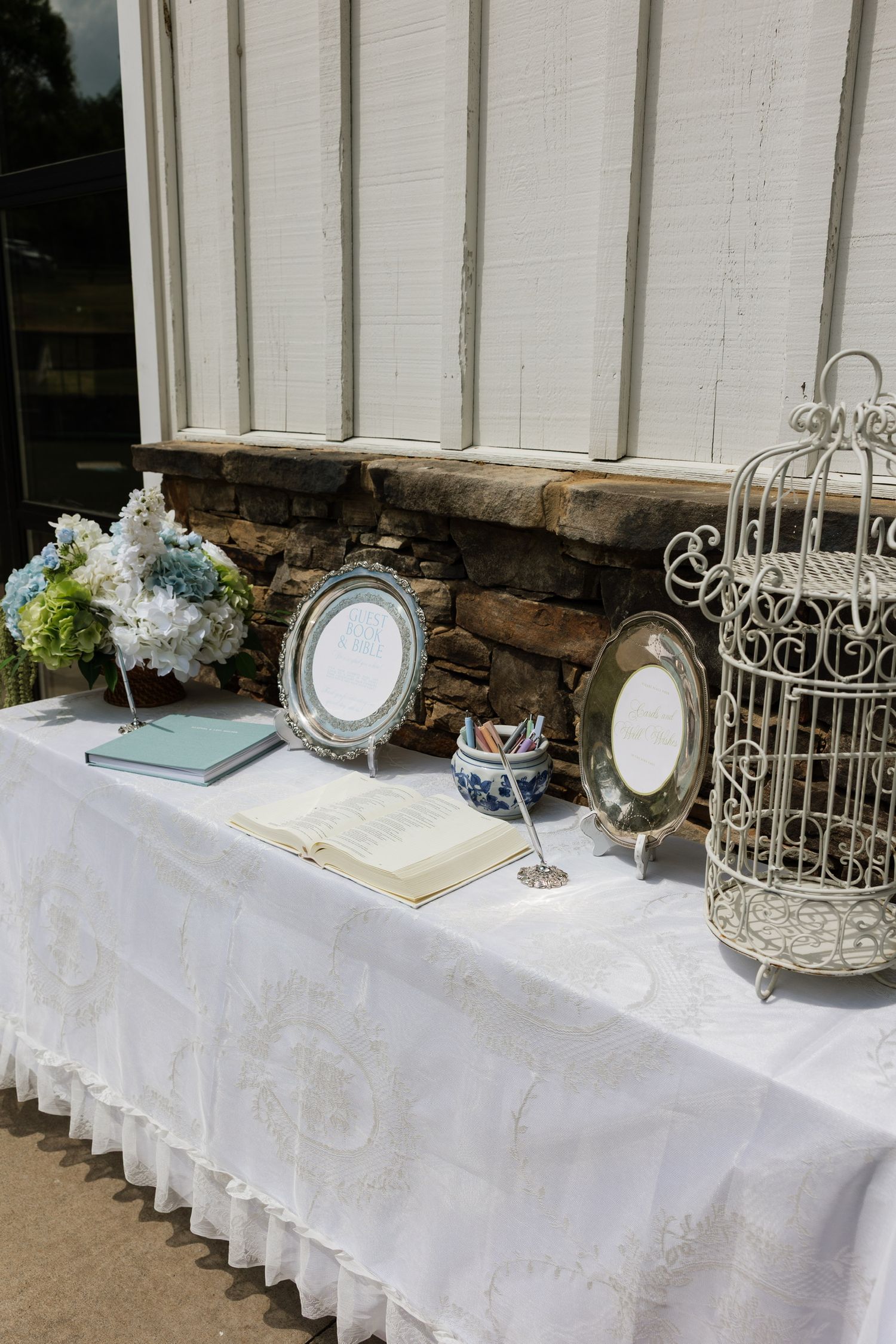 Wedding guest book table with silver frames, birdcage, hydrangea bouquet, and white lace tablecloth.