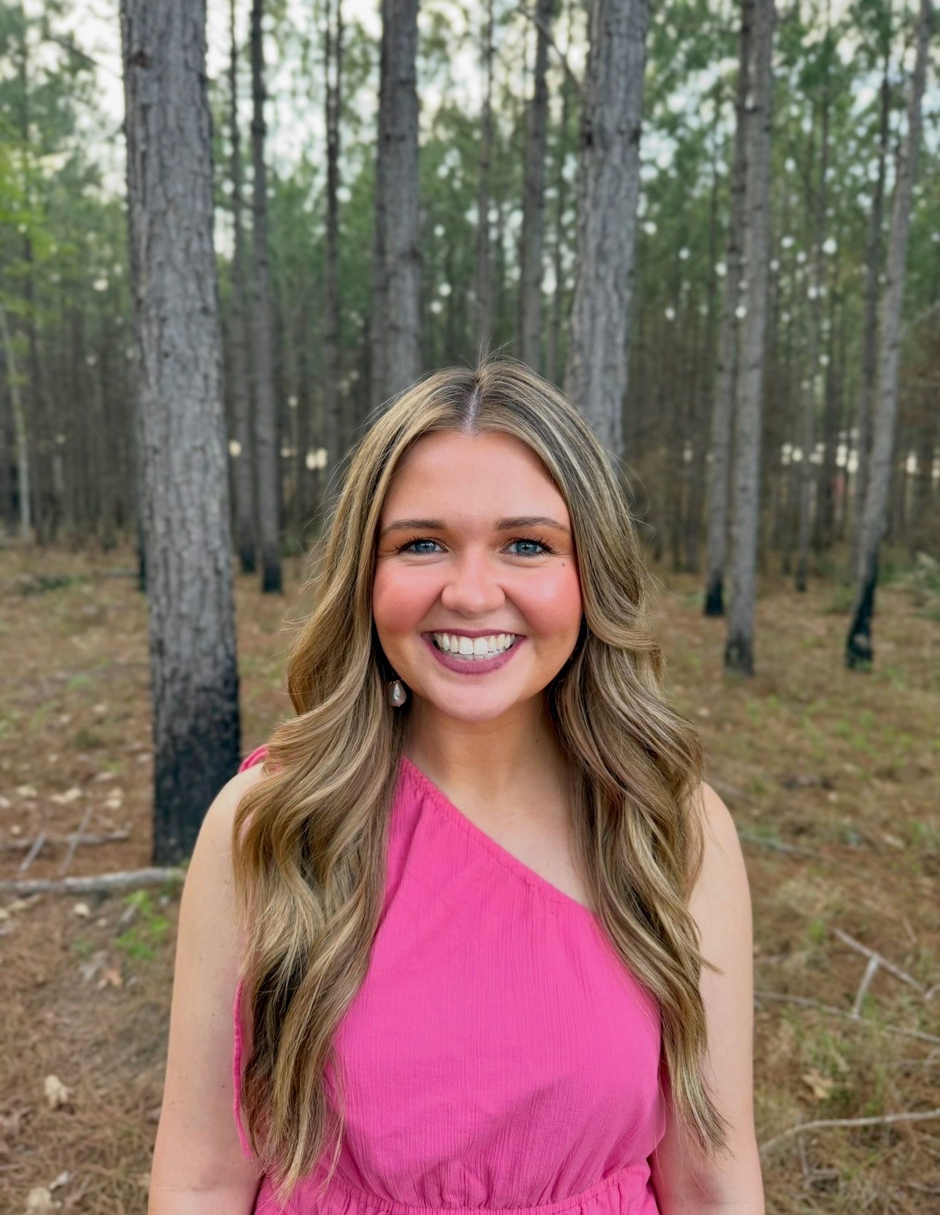 Smiling young woman in pink top with long blonde hair posing outdoors among tall pine trees.