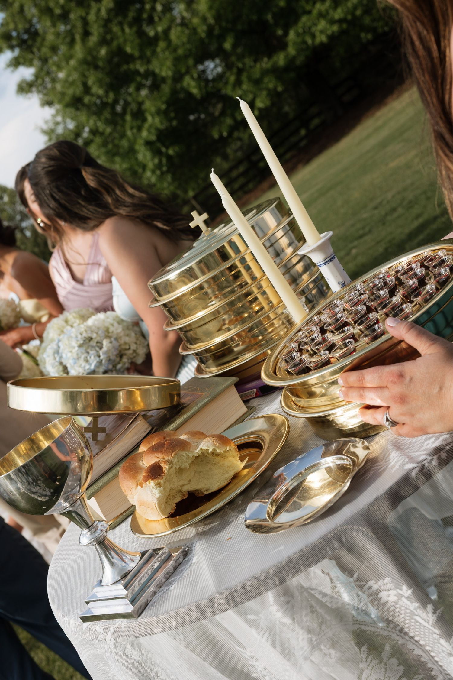 Outdoor communion table with gold chalices, candles, bread, and chocolates on white linen cloth during religious ceremony.