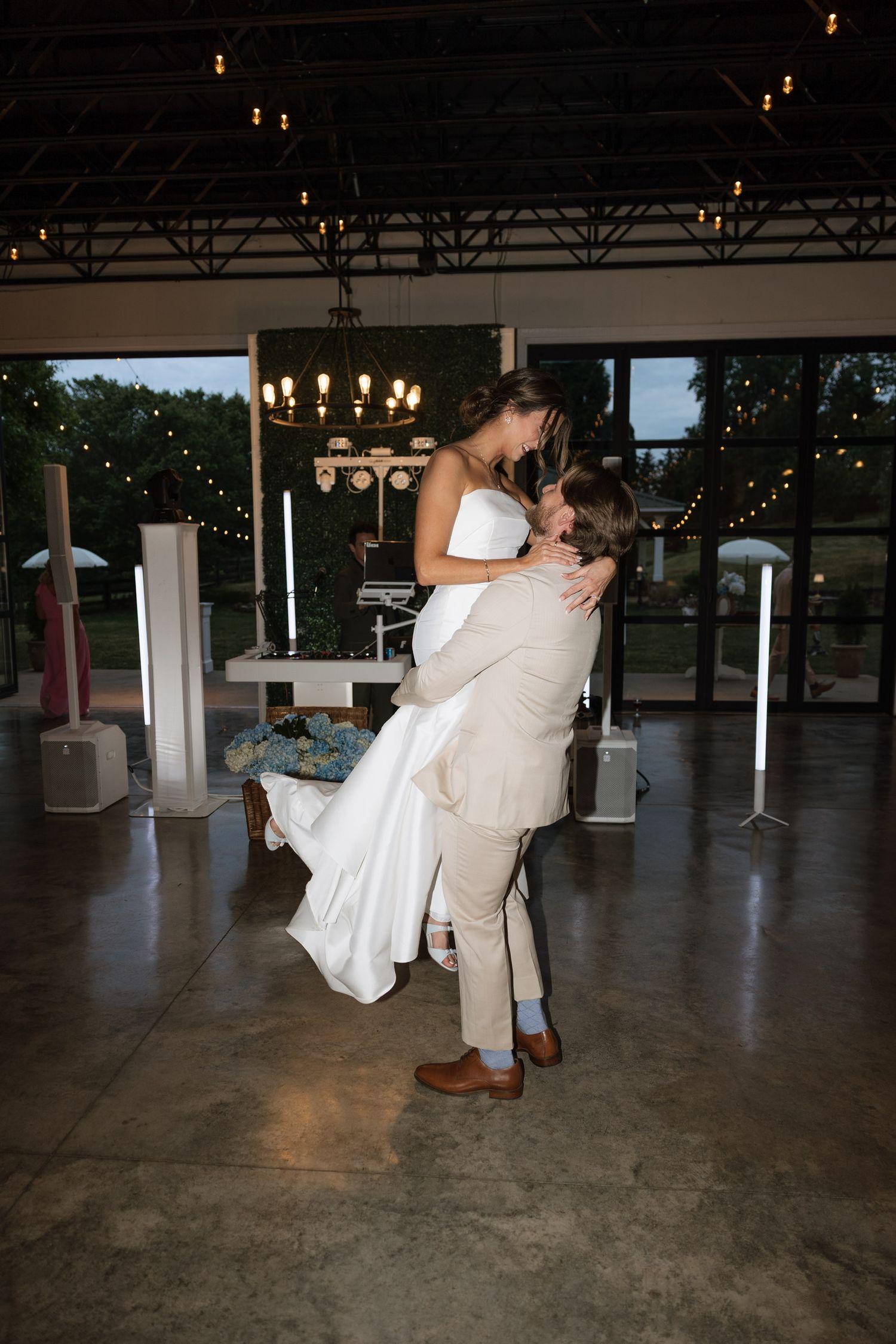 Groom lifts bride in white gown during first dance at elegant indoor wedding venue with string lights.