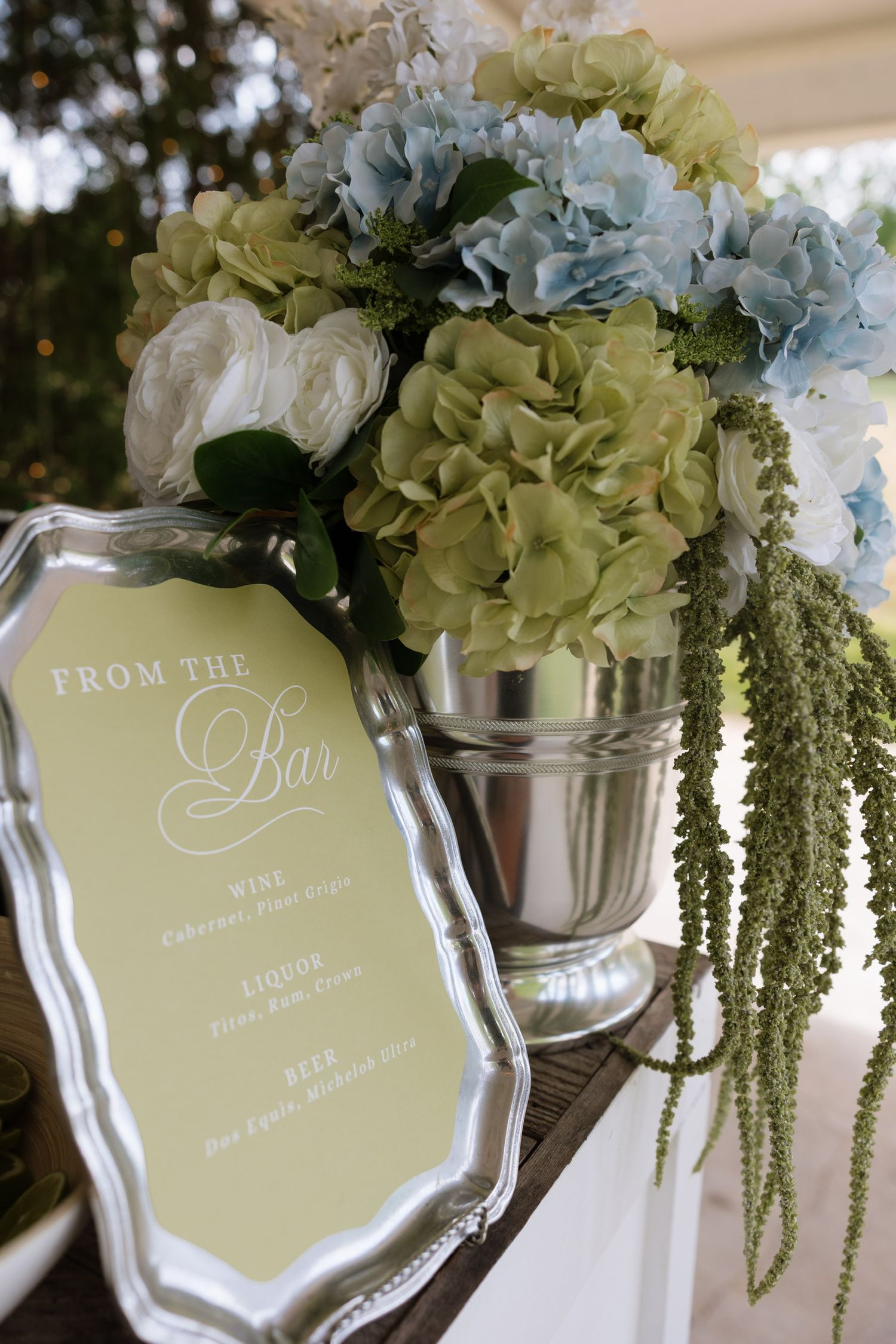 Silver tray bar menu sign beside lush blue and green hydrangea floral arrangement in silver bucket.