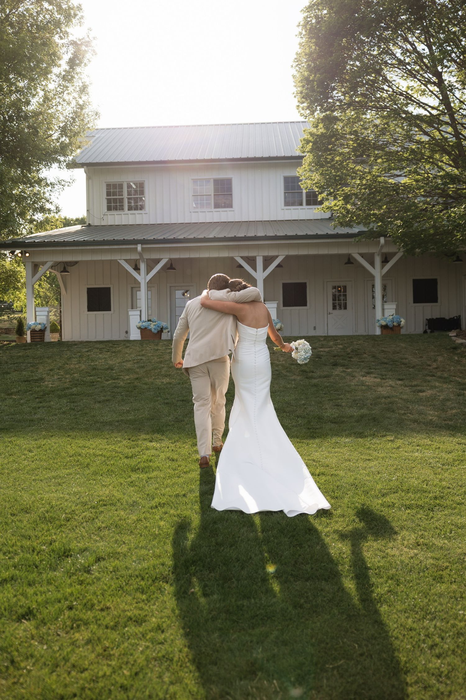 Bride and groom walking arm in arm across green lawn toward white farmhouse venue on sunny wedding day.