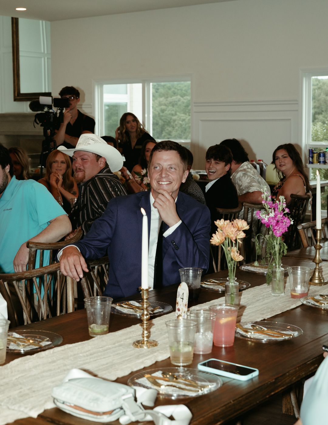 Wedding reception guests seated at long decorated table with floral centerpieces and candles.