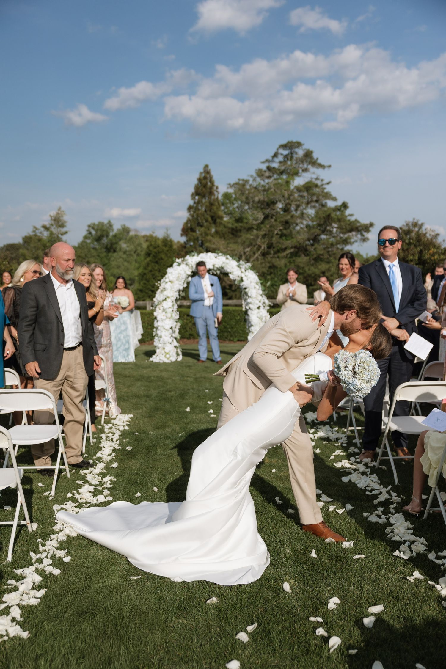 Groom dips bride for kiss while walking down outdoor aisle after wedding ceremony.