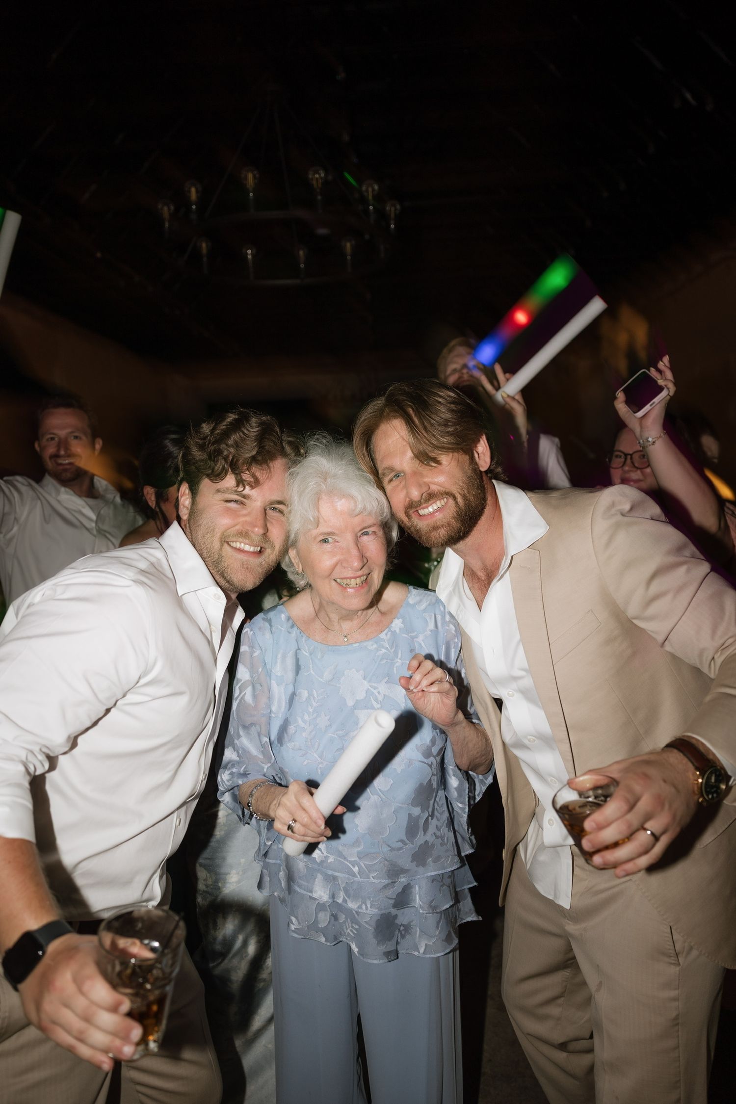 Two men in white suits pose with an elderly woman at a festive indoor party celebration.