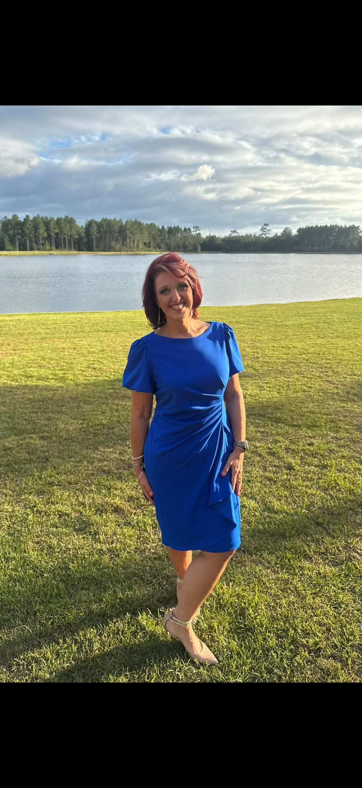 Woman in royal blue dress posing by a serene lake with lush green grass and pine trees in background.