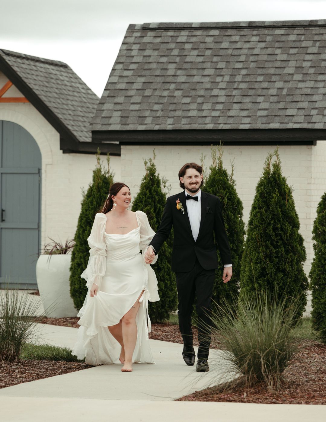 Bride and groom walking hand in hand outside white brick wedding venue with evergreen trees.