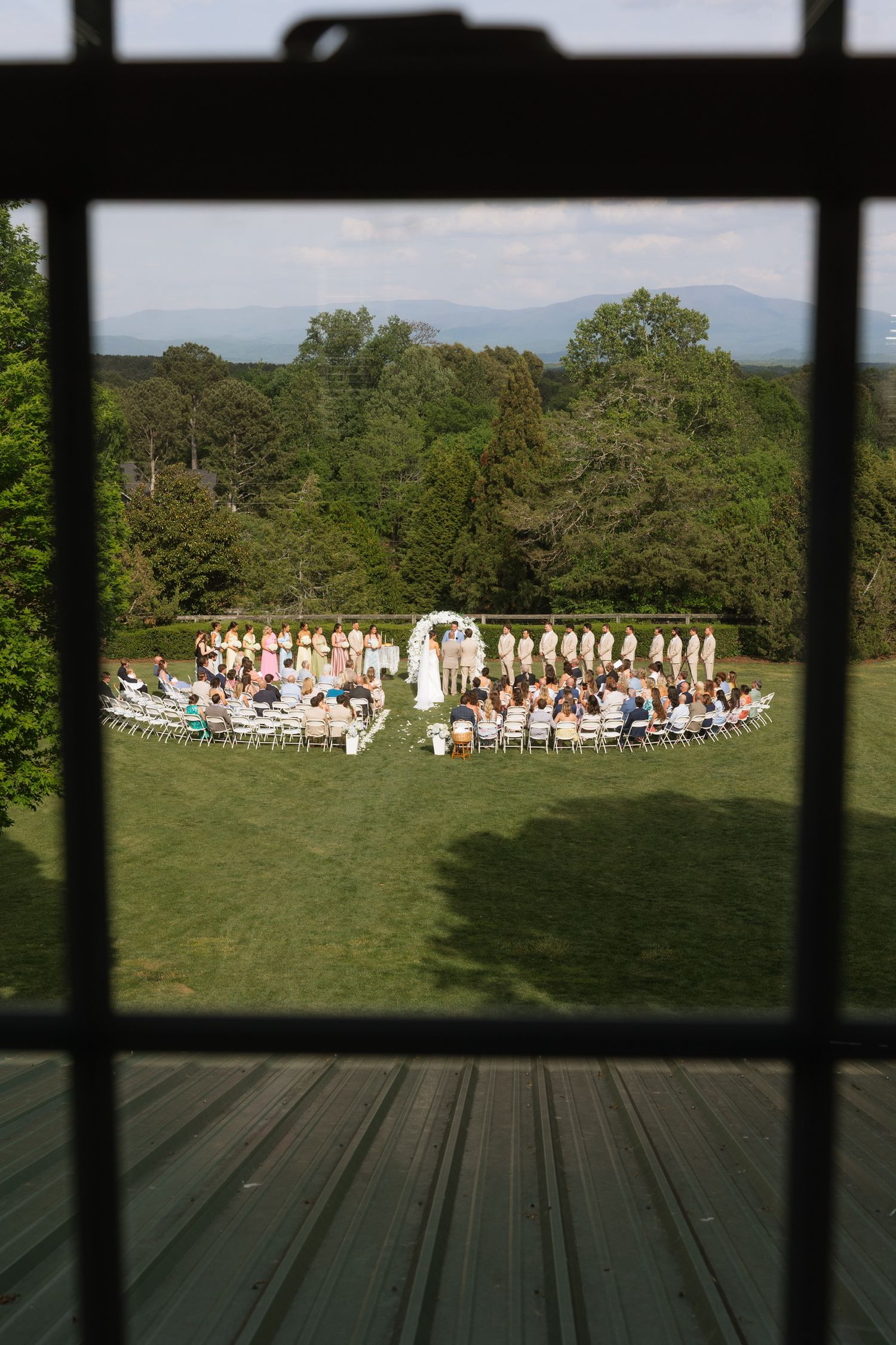 Outdoor wedding ceremony on green lawn with mountain backdrop, viewed through window, guests seated in semicircle.
