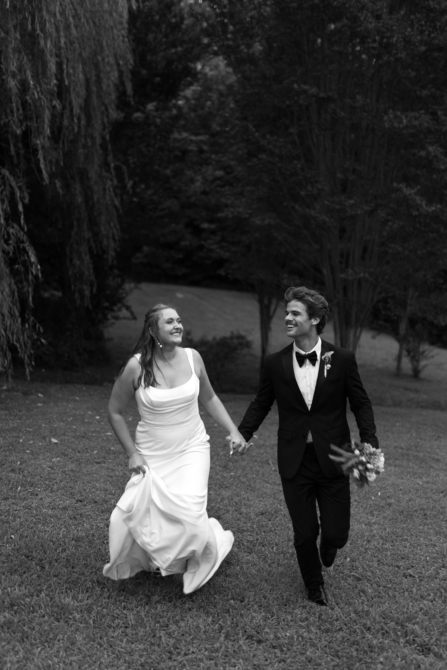 Black and white photo of smiling bride and groom holding hands while walking on grass with trees in background.