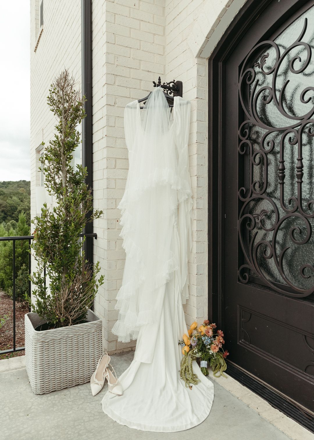 White wedding gown with veil hanging on iron door hook beside bridal shoes and bouquet.